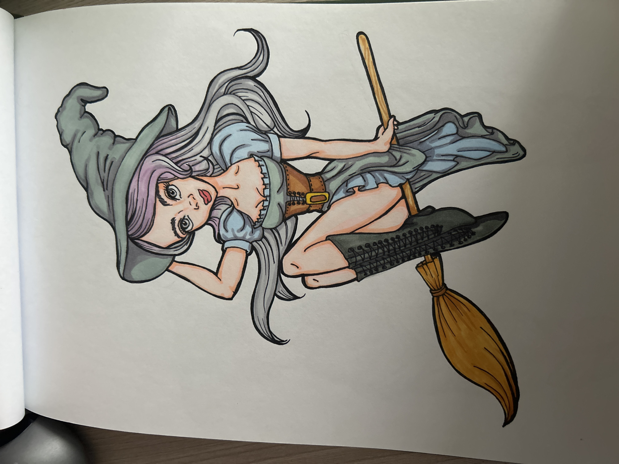 A drawing depicts a young woman with long, wavy, light purple and gray hair, wearing a pointy green witch's hat. She has large, expressive eyes, rosy cheeks, and red lips. Her attire includes a light blue ruffled top, a brown corset with a gold buckle, a flowing gray skirt, and tall black laced boots. She is posed sitting on a wooden broomstick, holding onto her hat with one hand. The drawing is colored with what appears to be markers or colored pencils. The background is plain white paper. The image is a close-up shot of the drawing, with the top left corner of a spiral-bound sketchbook visible. There is no indication of a city, location, or any elements that would suggest Angarsk, Russia.