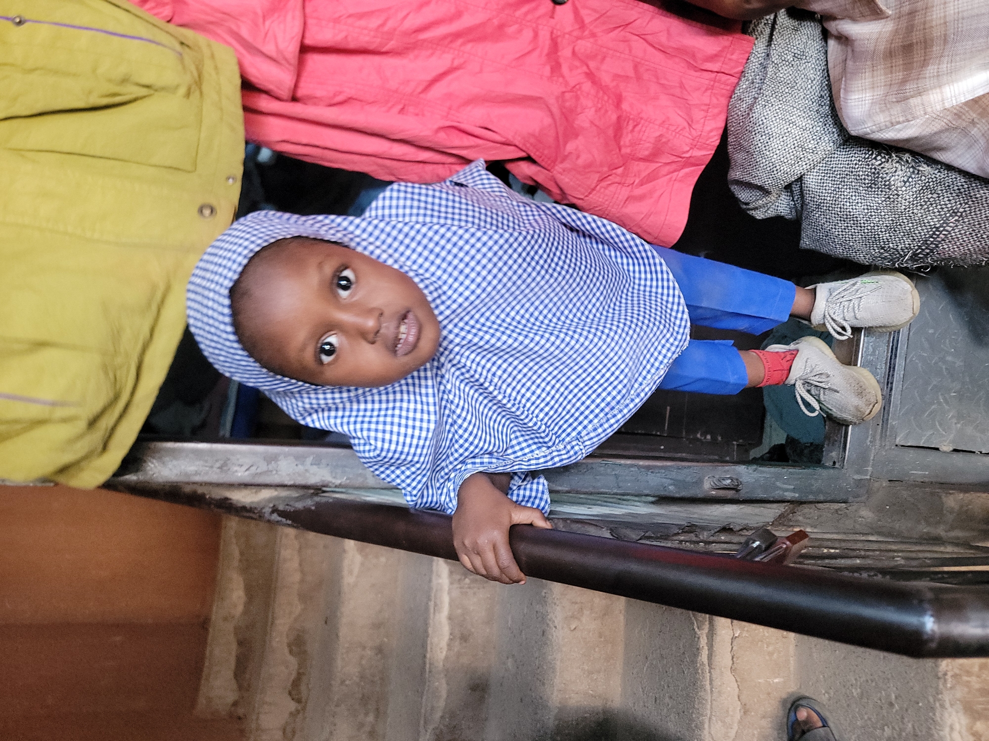 A young child with dark skin and short hair looks directly at the camera with a bright, inquisitive expression. The child is wearing a blue and white checkered top or dress and blue trousers. A small red cuff or band is visible on the child's right wrist (left side of the image). The child's feet are clad in worn white sneakers with laces.
The image is captured from a slightly upward angle and is rotated 90 degrees clockwise. If viewed upright, the child would appear to be leaning over a dark metal railing or bar, which they are grasping with their right hand.
The background is filled with various draped and hanging garments, suggesting a setting such as a market stall, a shop, or a storage area for clothes. Visible fabrics include a prominent yellow or lime green jacket, a bright pink or red fabric, and patterned grey and light brown textiles. Below the railing, there are concrete steps or flooring, and a darker, possibly metallic, surface behind the child's feet. Another person's foot, wearing a dark sandal or flip-flop, is partially visible at the bottom right.
The scene depicts a candid moment from daily life, with the child as the central subject. The lighting is soft and diffused, indicating an indoor or shaded outdoor environment in Maiburiji, Nigeria. No visible text is present in the image.