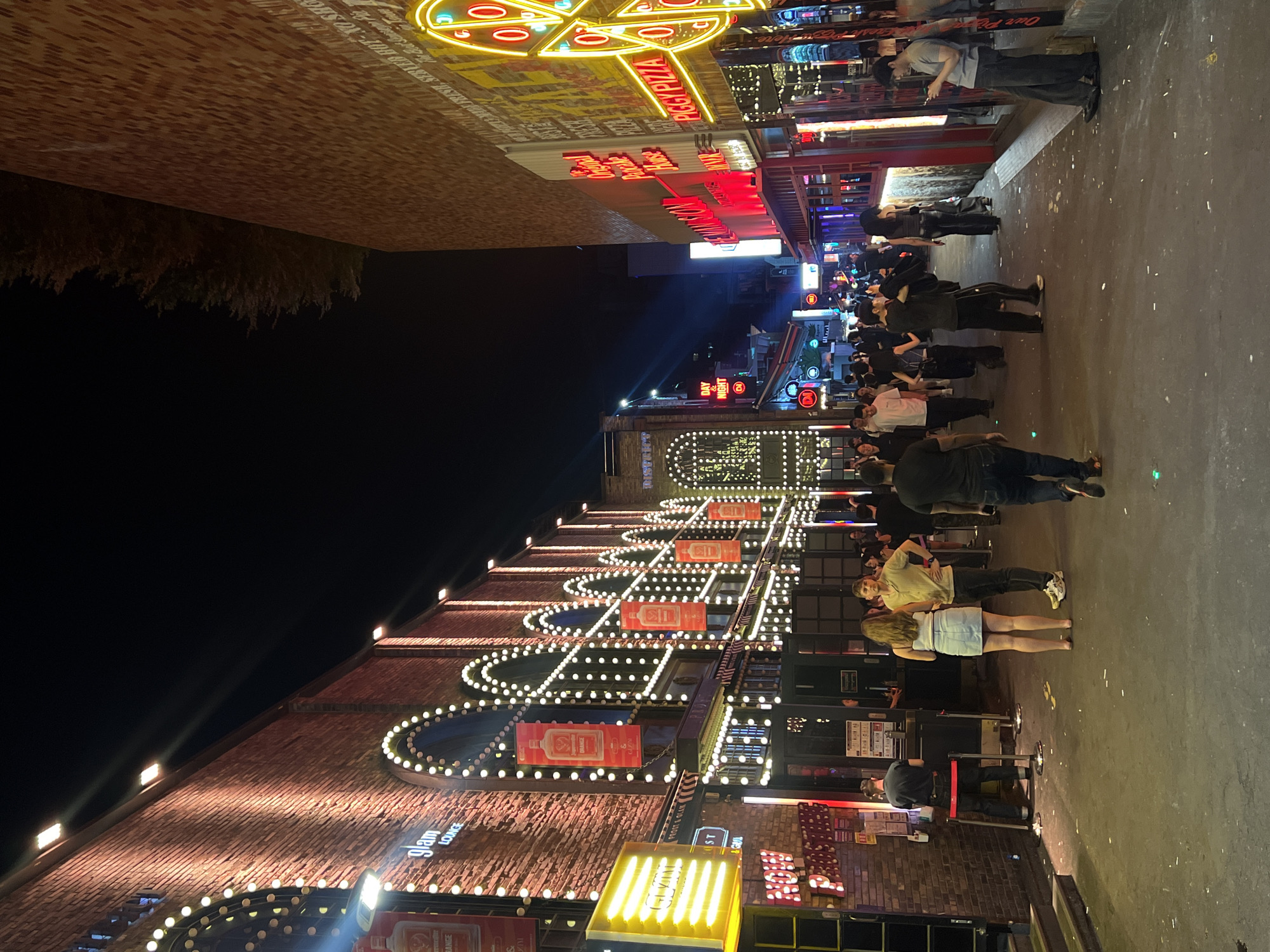 The image shows a bustling street scene in Seoul, South Korea, at night. A crowd of people is walking along the sidewalk past a row of buildings with brightly lit facades. The buildings are decorated with strings of lights and neon signs. The signs include the name "Peggy Pizza" and "Glam Lounge". The lighting is vibrant, creating a lively atmosphere. The sky is dark, and there is sparse greenery above the structures.