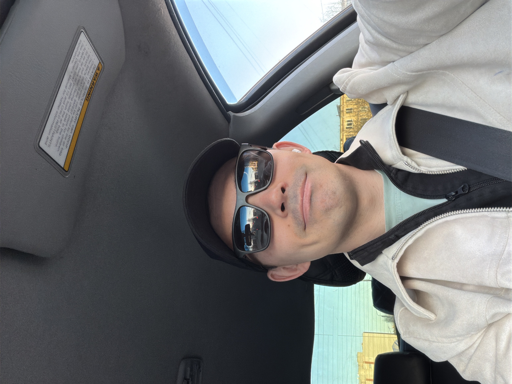 The image shows a selfie of a person inside a vehicle. The person is wearing sunglasses and a black cap. The reflection in the sunglasses reveals a street scene, suggesting the car is in motion. The person is also wearing a light-colored jacket over a darker layer and has a seatbelt fastened.
The interior of the car is visible, including the ceiling, a sun visor with a warning label, and a sunroof.
The location is possibly Izhevsk, Russia based on the reflection in the sunglasses.