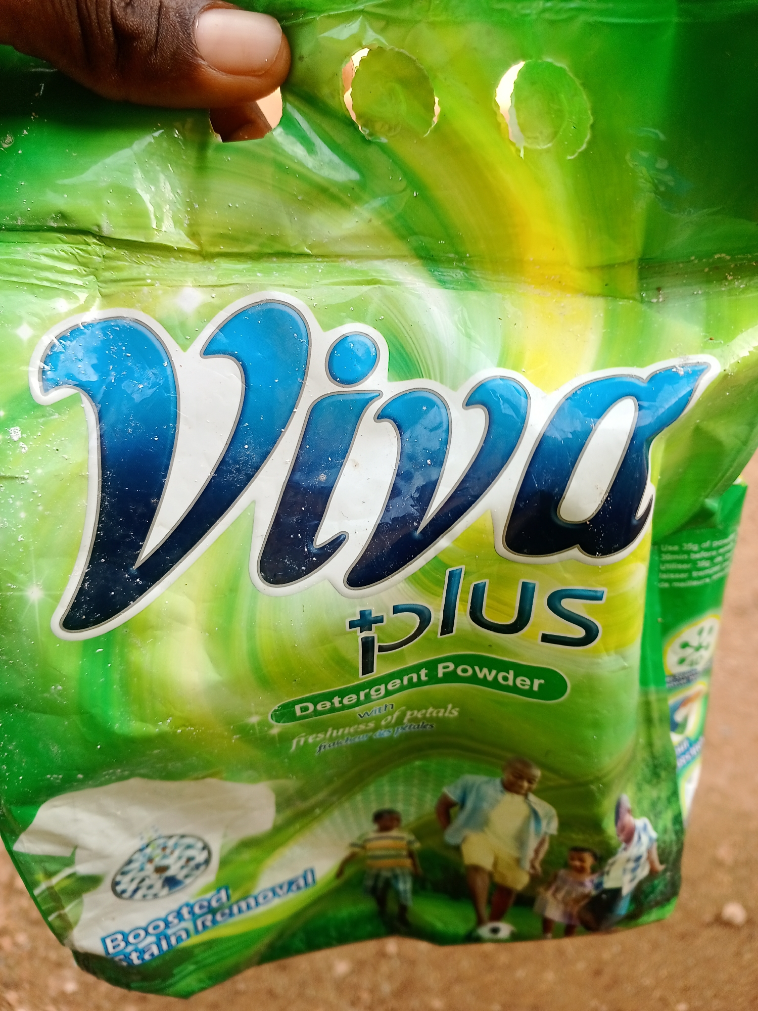 A hand holds a bag of Viva Plus Detergent Powder, the bag being primarily green with yellow accents. The branding "Viva plus" is prominently displayed in large blue lettering with a white outline. Below this, smaller green text reads "Detergent Powder" and then "with freshness of petals". Towards the bottom left of the bag, white text states "Boosted Stain Removal" above a circular graphic depicting a washing machine drum with water and suds. The bag is also adorned with an illustration of a family playing soccer in a grassy field, suggesting outdoor activity and a pleasant, family-oriented scene. The overall impression is a product designed for laundry care, emphasizing freshness and stain removal. A portion of another bag of the same product is visible to the right. The background consists of a reddish-brown, sandy surface, possibly the ground, and the lighting suggests it is daytime. A small amount of text in a smaller font is visible on the right side of the bag, which appears to be usage instructions in English and possibly French. The location is identified as Jalingo, Nigeria.