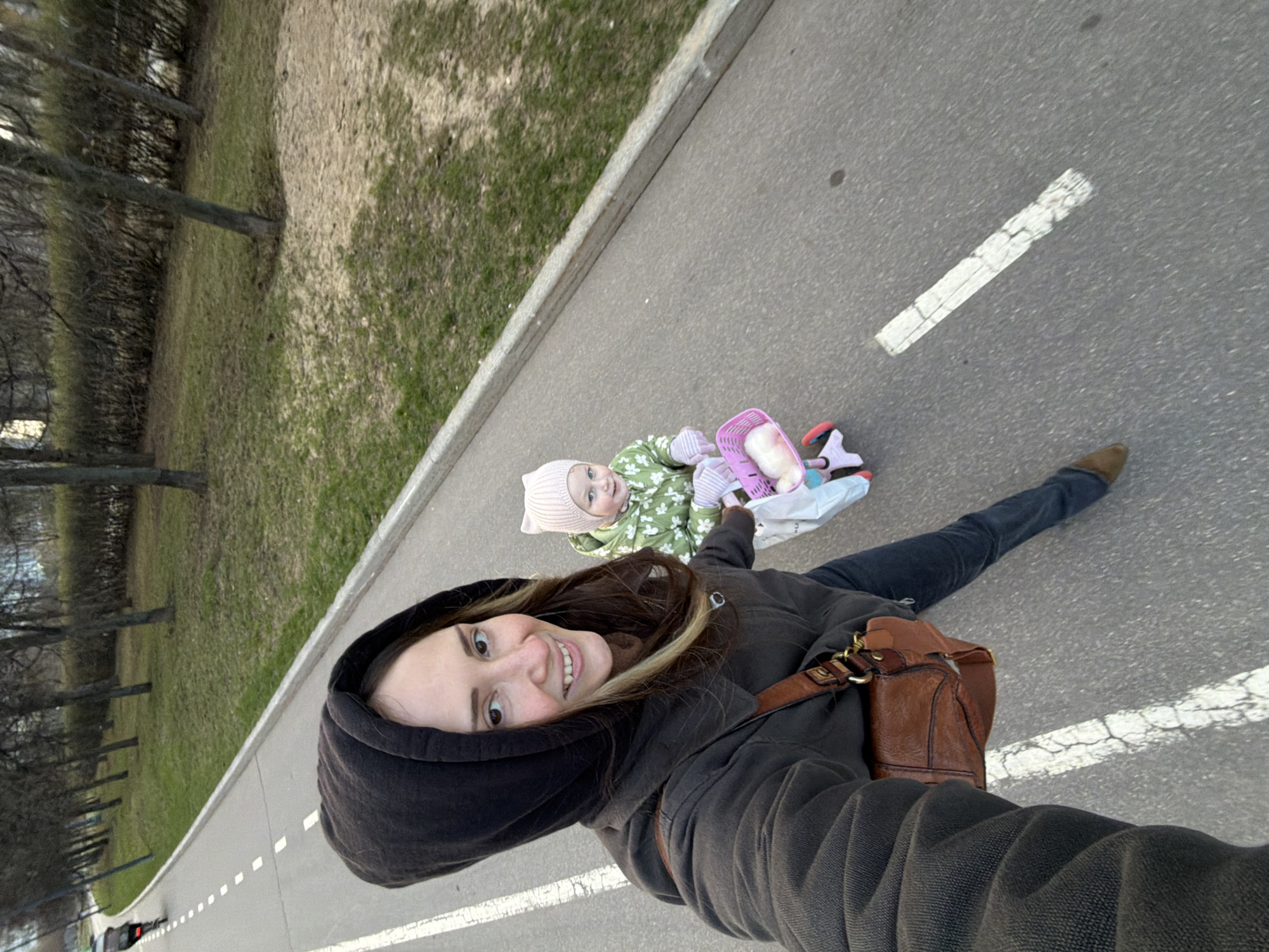 A woman in a dark hooded jacket and jeans is taking a selfie on a paved path. She has a brown leather shoulder bag. Beside her is a child, likely an infant, wearing a pink hat and a green floral-patterned outfit in a carriage. The path has white dashed lane markings. To the side of the path is a grassy area with bare trees, indicating it may be early spring or late autumn. Based on the location request, the photograph appears to be taken in a public park in Khimki, Russia.
