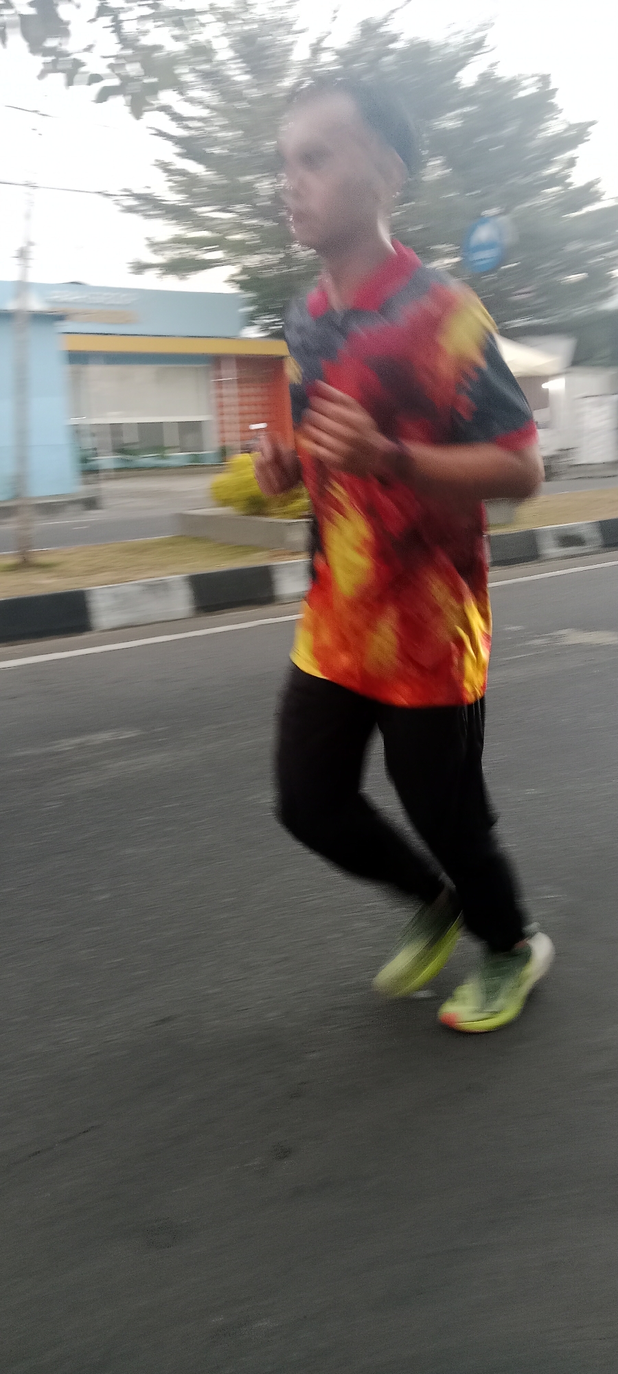 A low-angle, motion-blurred, full-body shot captures a person running outdoors. The subject, wearing a brightly colored, tie-dye t-shirt in shades of red, yellow, and black, along with black leggings, is in mid-stride. Their athletic shoes are a vibrant neon yellow and green. The running takes place on a paved road, with a curb and a grassy area visible to the left. In the background, a light blue building with orange trim and windows stands, suggesting an urban or suburban environment. The foliage of trees is present in the upper portion of the frame. The lighting indicates it is daytime, likely with overcast conditions given the diffused light. The specific location is identified as Ngemplak, Indonesia.