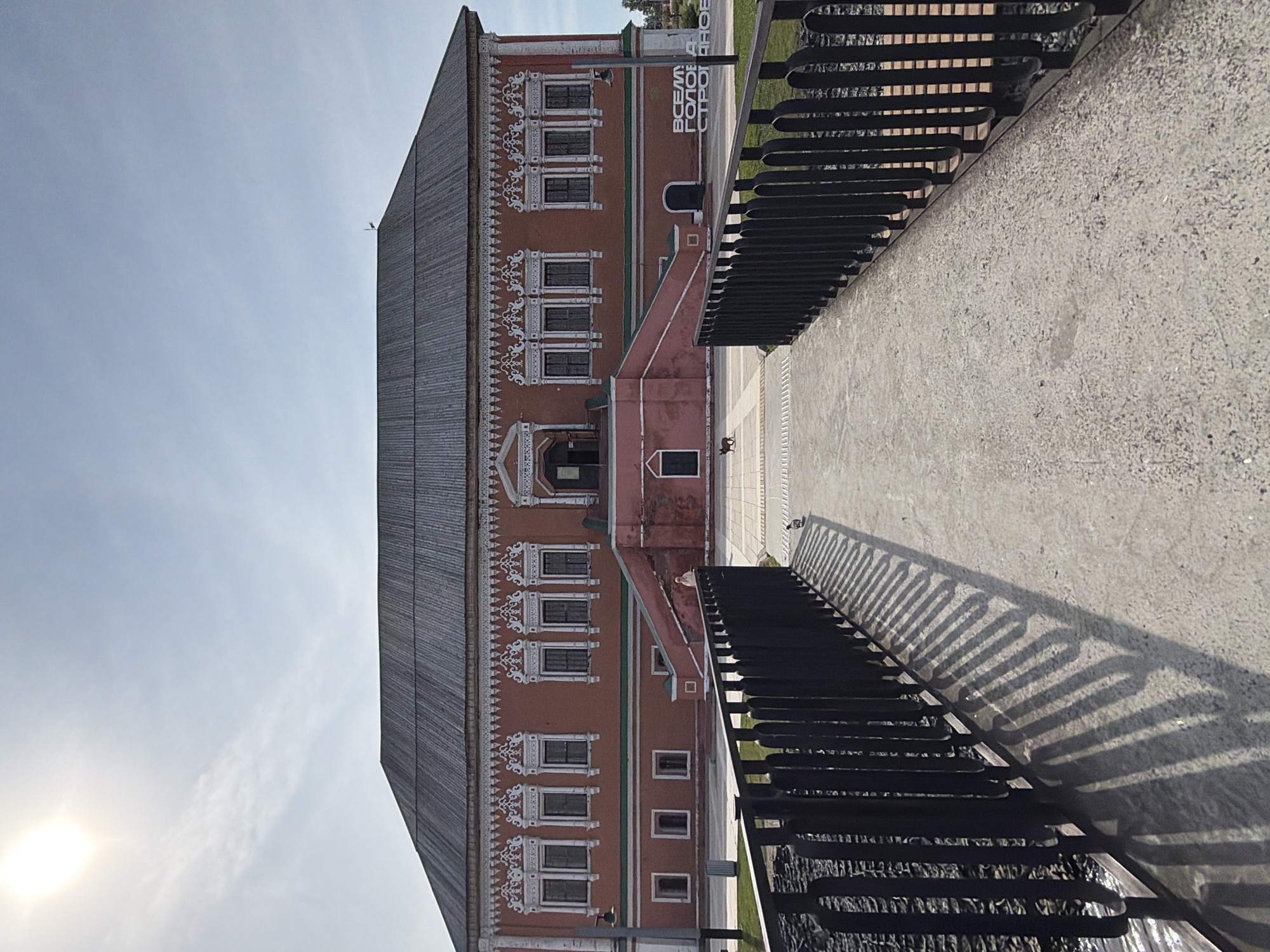 In the city of Usolye, Russia, a historical stone building with a dark, sloped roof stands under a bright, sunlit sky with some wispy clouds. The building's facade is predominantly a muted reddish-pink, adorned with intricate white decorative elements around numerous windows on the upper level. A grand staircase with pink risers leads up to the central entrance. In front of the building, a black metal fence borders a pathway that extends towards the viewer. A narrow body of water with dark, textured stones is visible beyond the fence on the left. A small, dark-colored dog-like animal walks on the paved area in front of the building. In the far right of the image, white text is visible on the building's wall. The foreground shows a concrete path leading up to the building, with the shadow of the fence cast across it.