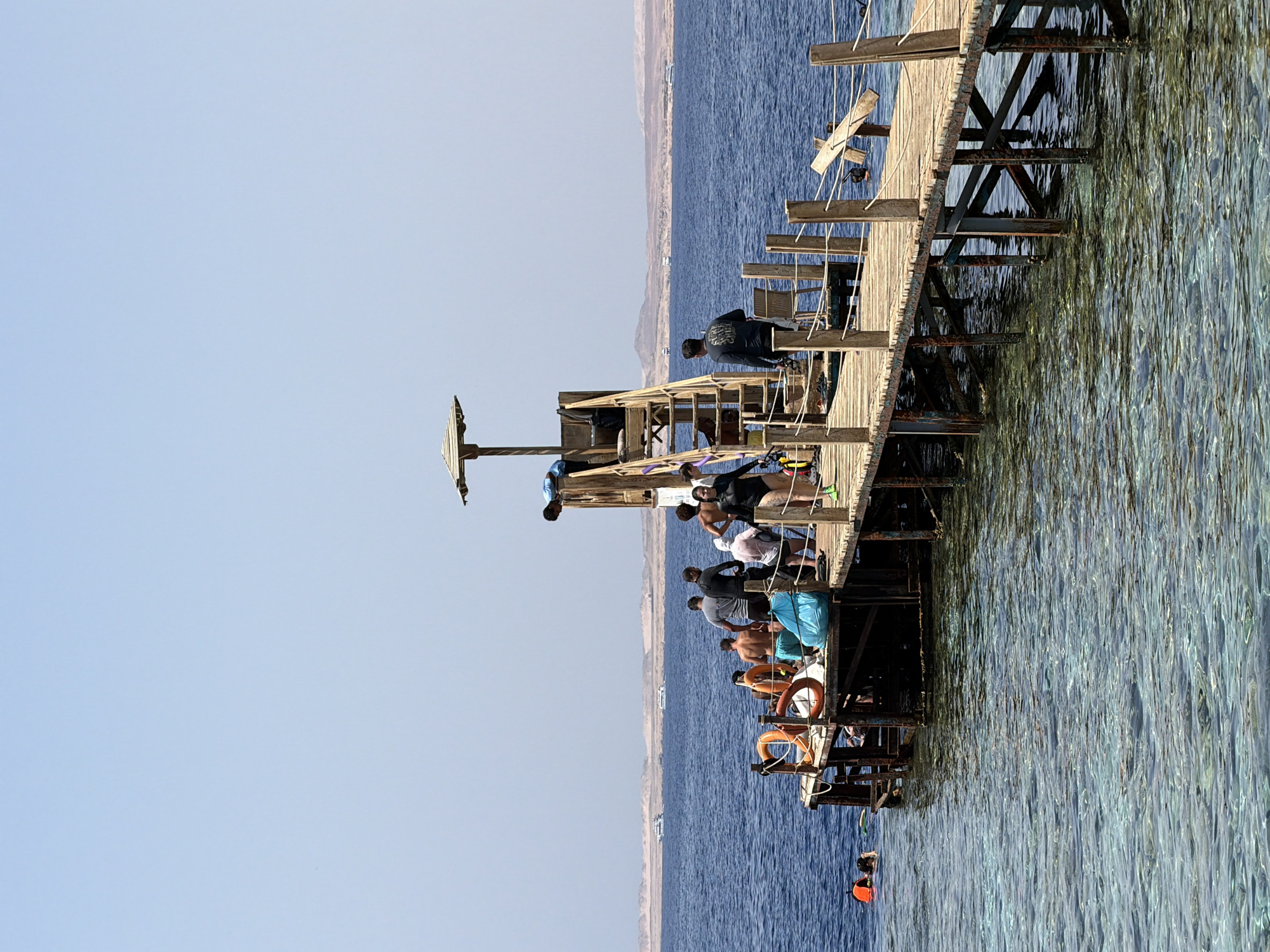 A wooden pier extends over clear, shallow water in Sharm El Sheikh, Egypt. Several people are on the pier, some wearing swimsuits and others casual clothing. One person is wearing a black wetsuit and appears to be preparing to enter the water, while others are observing the water or conversing. On the left side of the pier, two individuals are in the water, wearing orange life vests and snorkeling gear. A lifeguard tower with a thatched roof is situated at the end of the pier, and a person in a blue shirt is visible in the tower. The background shows a body of calm, blue water, a distant sandy shoreline with mountains, and several boats on the water. The sky is a clear, pale blue.
