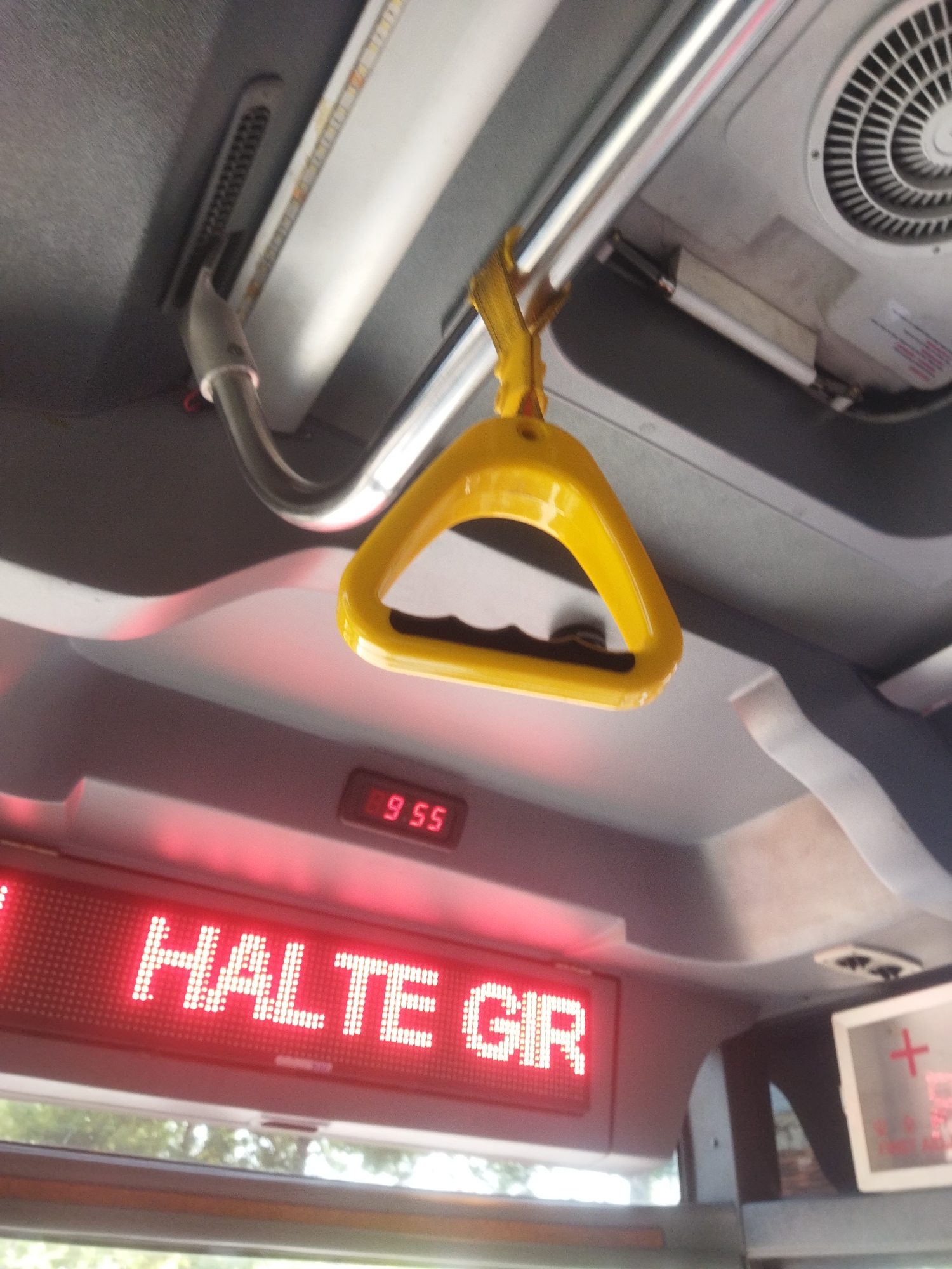 The image captures a close-up, interior view of a bus or public transport vehicle. In the foreground, a bright yellow plastic handle, designed for passengers to hold onto, is prominently displayed, hanging from a silver metal pole. Below the handle, a red LED display shows the text "HALTE GIR" in white, indicating a stop or destination. Above this display, a smaller red LED reads "9 55", likely representing the time. The ceiling of the vehicle is visible, with a grey textured surface, a ventilation grille on the right, and a strip light running along the top. The overall impression is one of a functional interior of a moving vehicle, likely in operation. The location of Wonokarto, Indonesia is not determinable from the image.