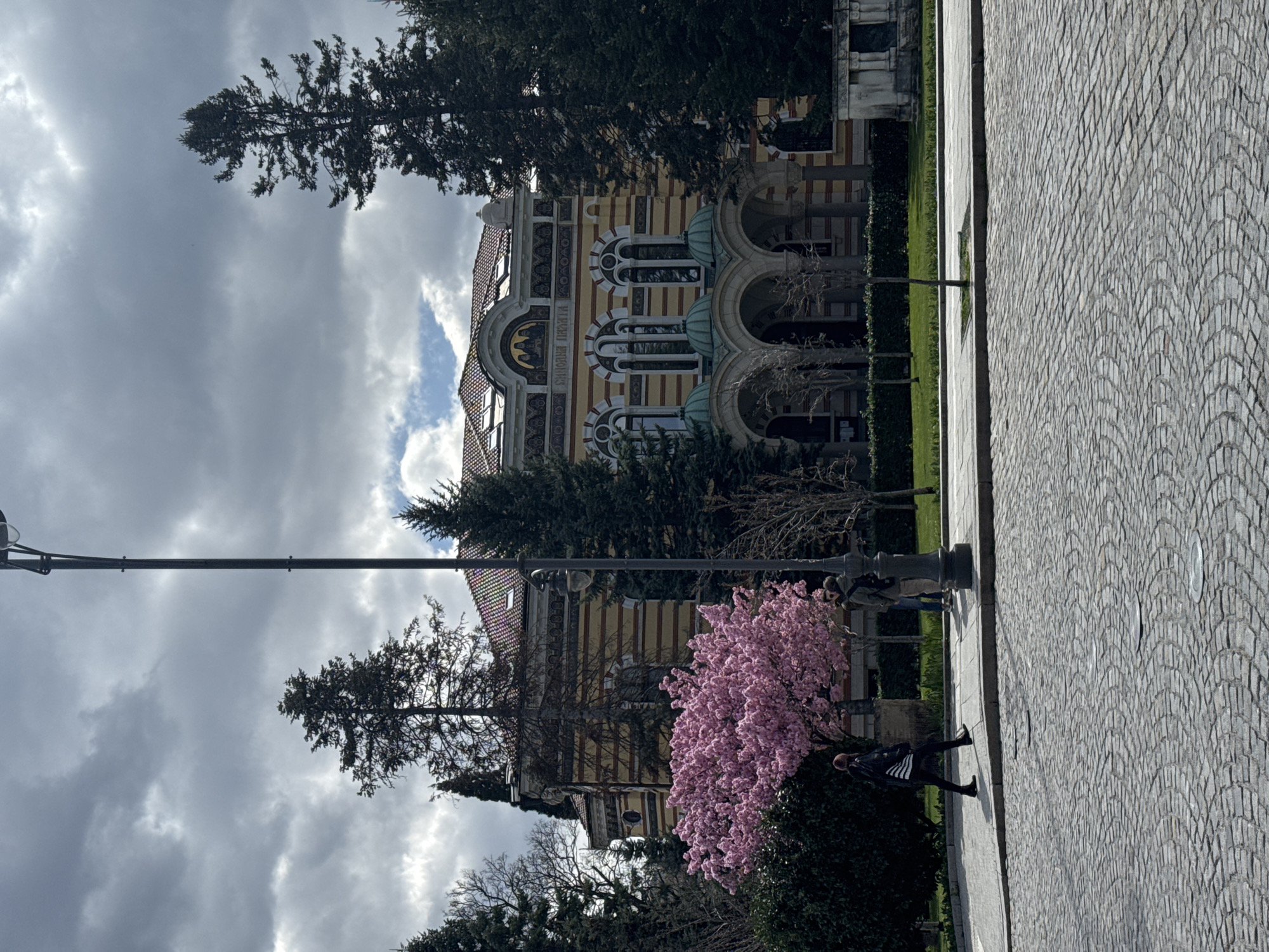 The image depicts a view in Sofia, Bulgaria, featuring the Sofia Mineral Baths building. The building has a distinctive architectural style with arched windows, yellow and white striped facades, and green domes. Tall trees partially obscure the structure. A pink blooming tree is in the foreground. A streetlight extends across the frame. On the right side of the frame, a paved walkway made of cobblestones is visible. A person is walking on the pavement. The sky above is filled with grey clouds.