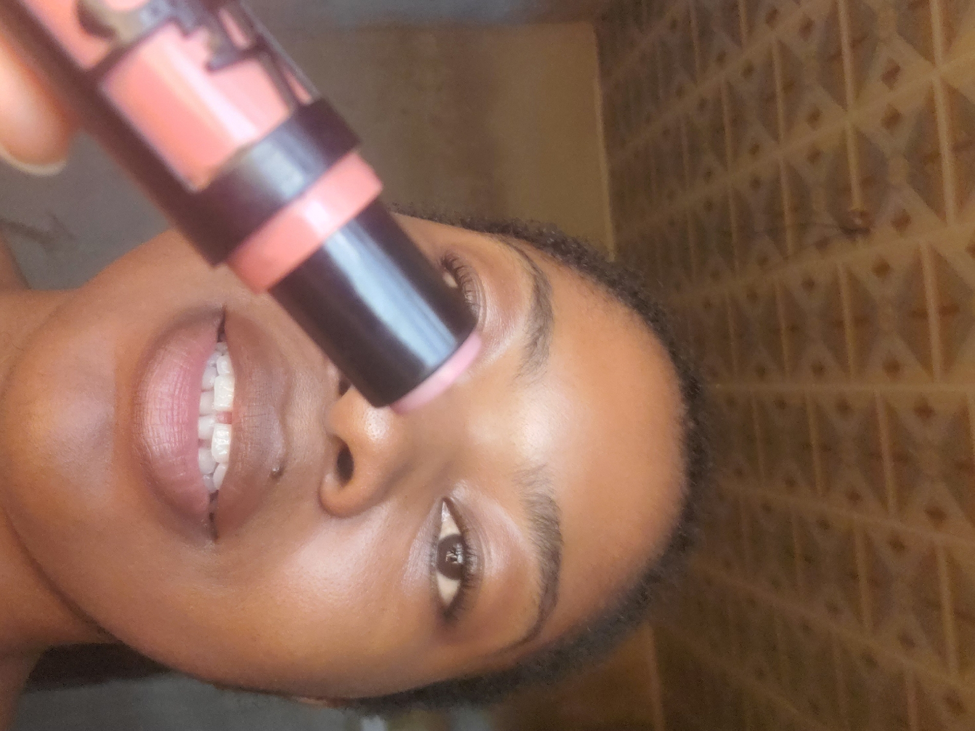 The scene is depicted in Kakau, Nigeria. A close-up shot features an individual, likely female, with dark skin and dark, short, curly hair framing the forehead. The individual's dark brown eyes are open, with visible eyelashes, and their gaze is directed towards the viewer. A small, silver piercing is visible on the right side of the upper lip. The mouth is slightly open, revealing white teeth with a distinct gap between the two central incisors. The individual holds a black and coral-pink makeup stick in the foreground, with the light pink product tip extended. The stick is positioned diagonally across the lower-left portion of the face, obscuring part of the lower jaw and neck. The background consists of a light brown, patterned ceiling with a grid of raised squares or diamonds in the upper half of the frame. Below the ceiling, a plain, light brown wall is visible, out of focus.