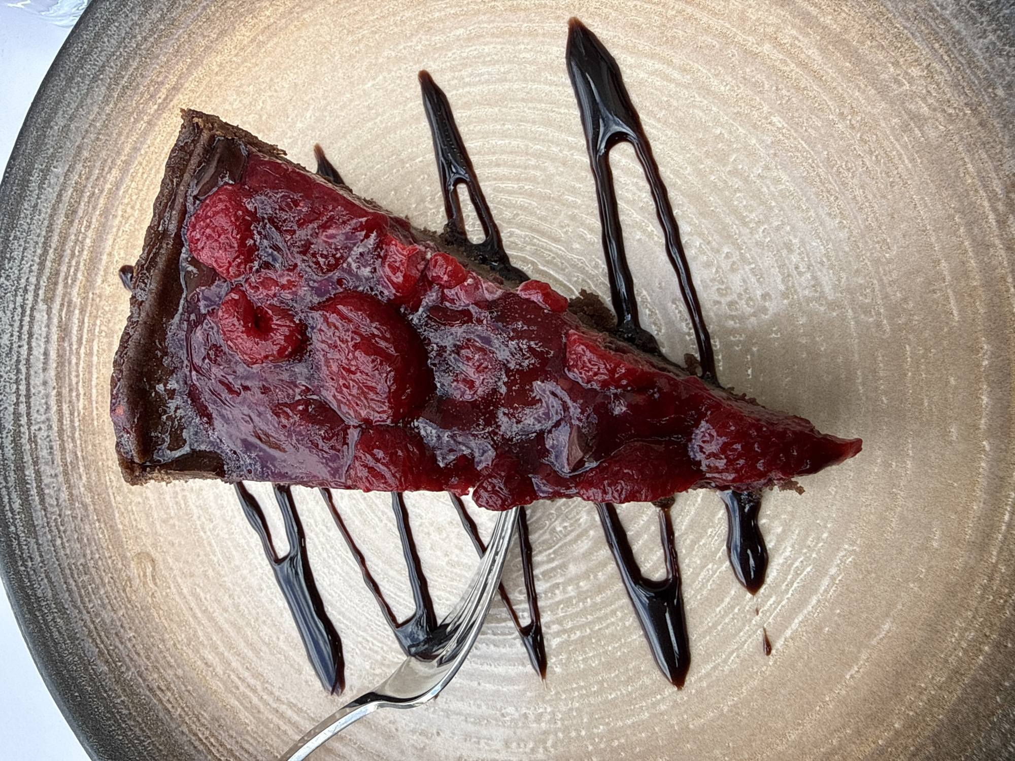 A close-up, high-angle shot shows a slice of dark chocolate tart topped with glazed red raspberries, resting on a textured, round plate. The plate has a light tan center with circular striations and a dark gray rim. Drizzles of dark chocolate sauce adorn the tart and plate, some pooling at the base. A silver fork is positioned near the bottom of the tart. The image appears to be taken in a well-lit setting. Varna, Bulgaria is not apparent in this image.