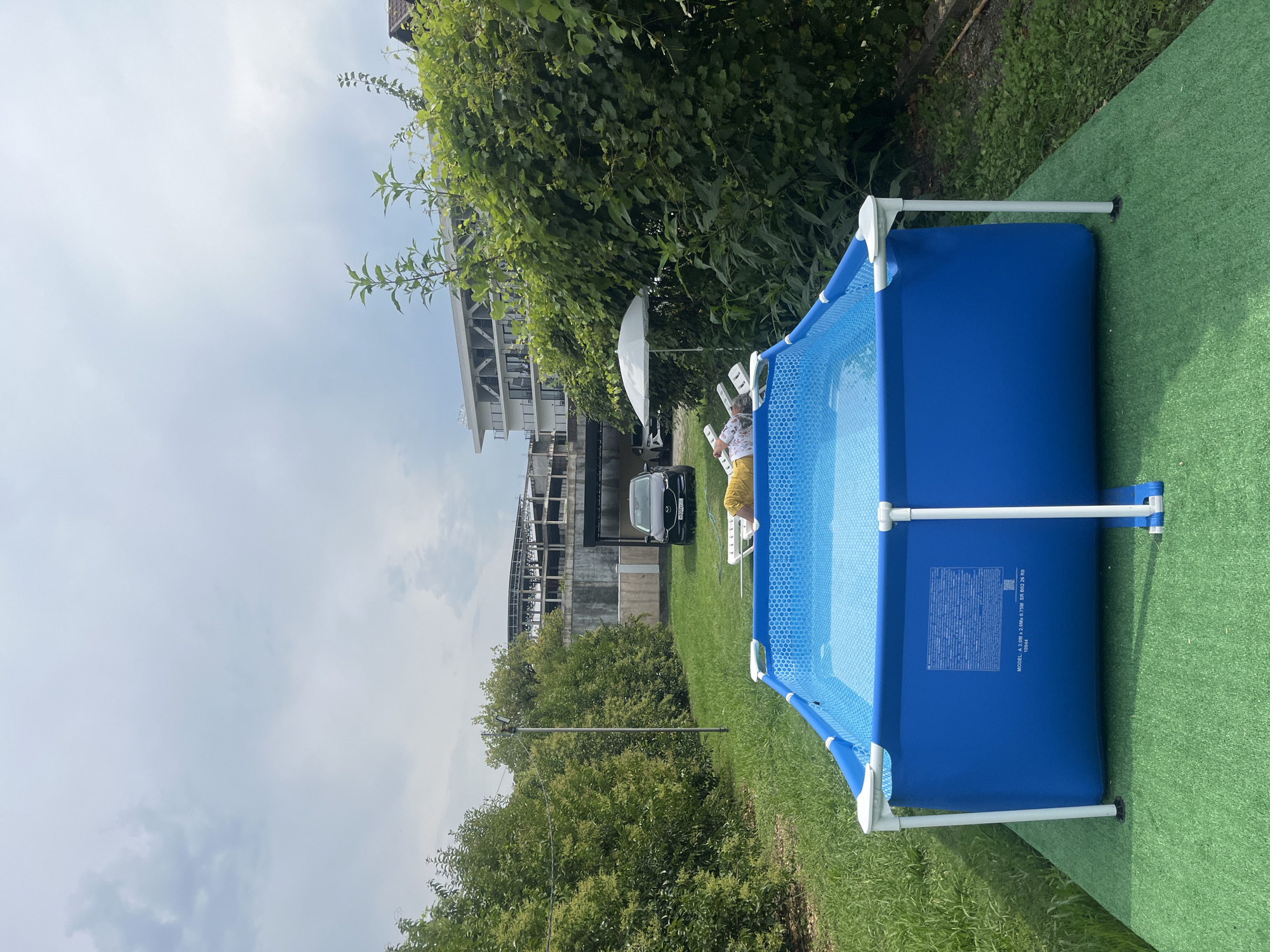 A person is relaxing on a lounge chair next to a blue, rectangular above-ground swimming pool in a grassy yard. The pool is filled with water and has a hexagonal pattern on the bottom. A white umbrella is set up next to the lounge chair, providing shade. In the background, there is a dark-colored car parked on a paved area, and beyond that, a multi-story building under construction. Lush green trees and bushes surround the yard, and a cloudy sky is visible overhead. The scene appears to be a residential backyard in Sokhumi, Abkhazia.