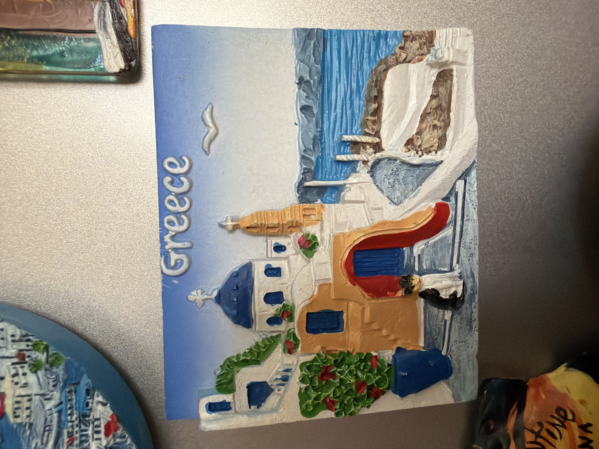 The image is a close-up photograph of a souvenir magnet depicting a scene from Greece. The magnet features white buildings with blue domes and accents, characteristic of Greek architecture. There is a prominent blue ocean in the background with distant islands. A small black and white cat is positioned on the cobblestone path in the foreground. The word "Greece" is written in raised, white lettering at the top of the magnet. The magnet is attached to a metallic surface, likely a refrigerator. The photograph does not depict Pazardzhik, Bulgaria.