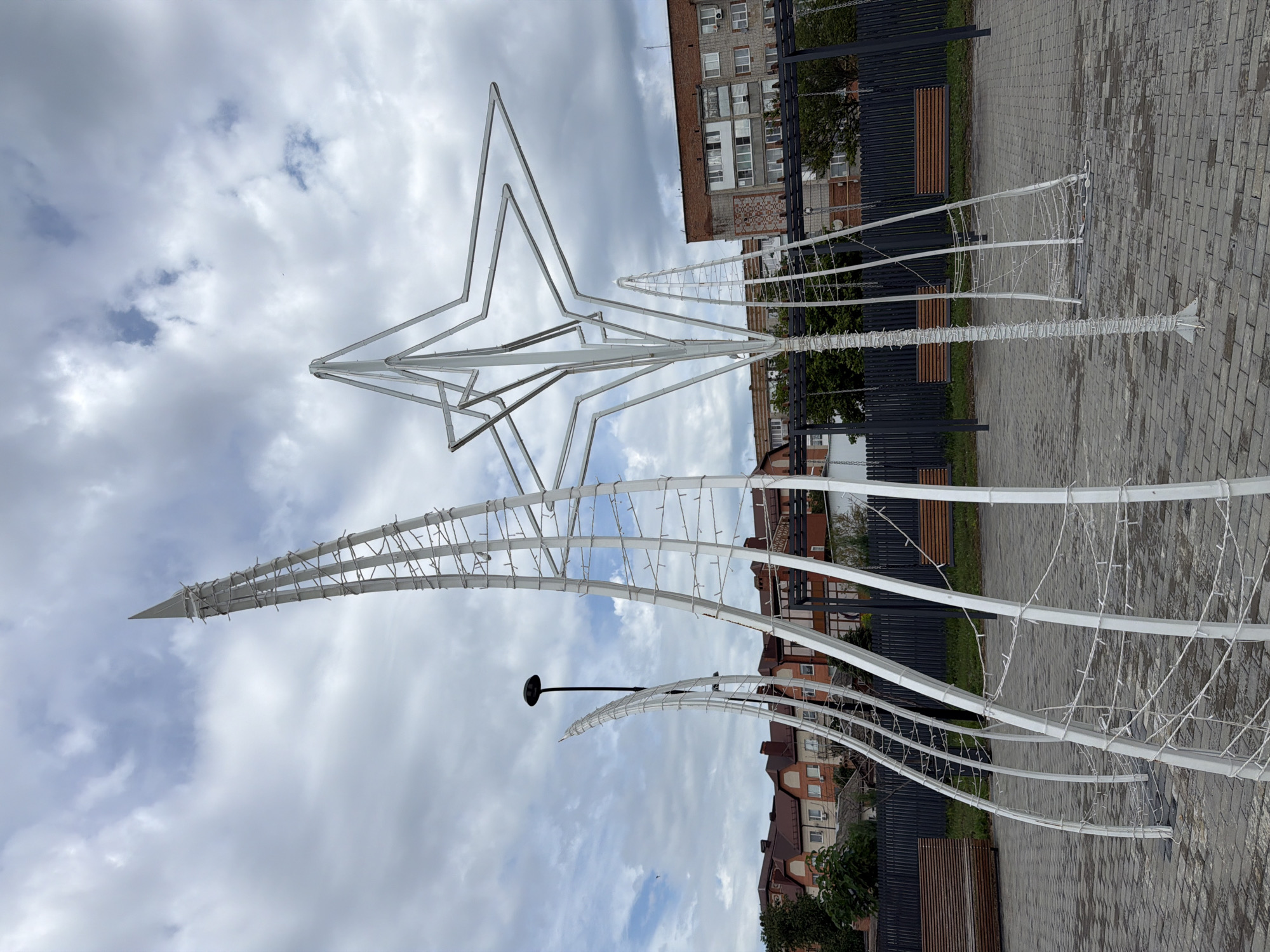 The image depicts an outdoor scene in Leningradskaya, Russia, featuring a large, white, abstract metal sculpture resembling a star and a towering spike. The sculpture is adorned with string lights. In the background, residential buildings and trees are visible. The ground is paved with gray bricks, and there are benches and a lamppost. The sky is cloudy.