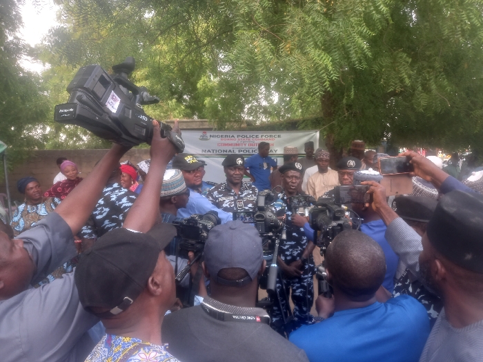 The image shows a crowd of people gathered in Maiduguri, Nigeria (latitude: 11.867, longitude: 13.123). The crowd is gathered around a group of men who appear to be police officers. There are many reporters and photographers in the crowd. The image is taken from the perspective of someone in the crowd, looking toward the police officers. Some people are holding cameras, cell phones, and other recording devices. There are also some women and children in the crowd. The image was taken in front of a wall with a banner reading "NIGERIA POLICE FORCE POLICE COMMUNITY OUTREACH NATIONAL POLICE DAY". The officers are wearing blue uniforms and have dark-blue berets. There are various people taking pictures of the officers with their phones and cameras. The scene is busy and chaotic, but there are no obvious signs of violence or aggression. The surrounding environment is a combination of greenery, some trees and a wall behind the group of people.