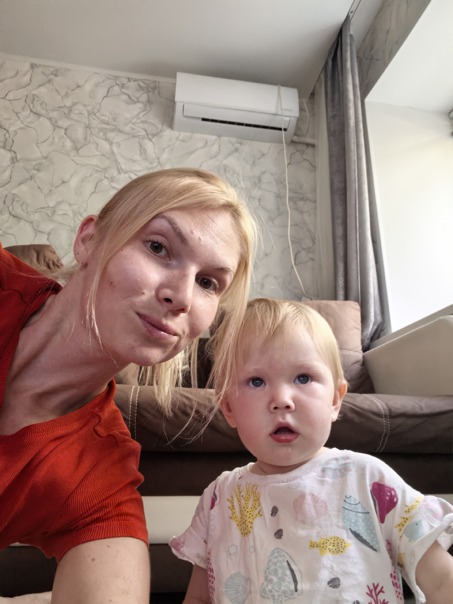 A selfie shows a fair-skinned woman with blonde hair and a toddler with blonde hair in a living room. The woman, wearing a red shirt, is on the left, looking at the camera with a slight smile. The toddler, in a white shirt with a shell pattern, is on the right, looking up with a surprised expression. Behind them, a brown sofa is visible, and a window with grey curtains is on the right. An air conditioner is mounted on a wall with marbled wallpaper. The lighting suggests it is daytime. The scene is likely indoors, in a residential setting.