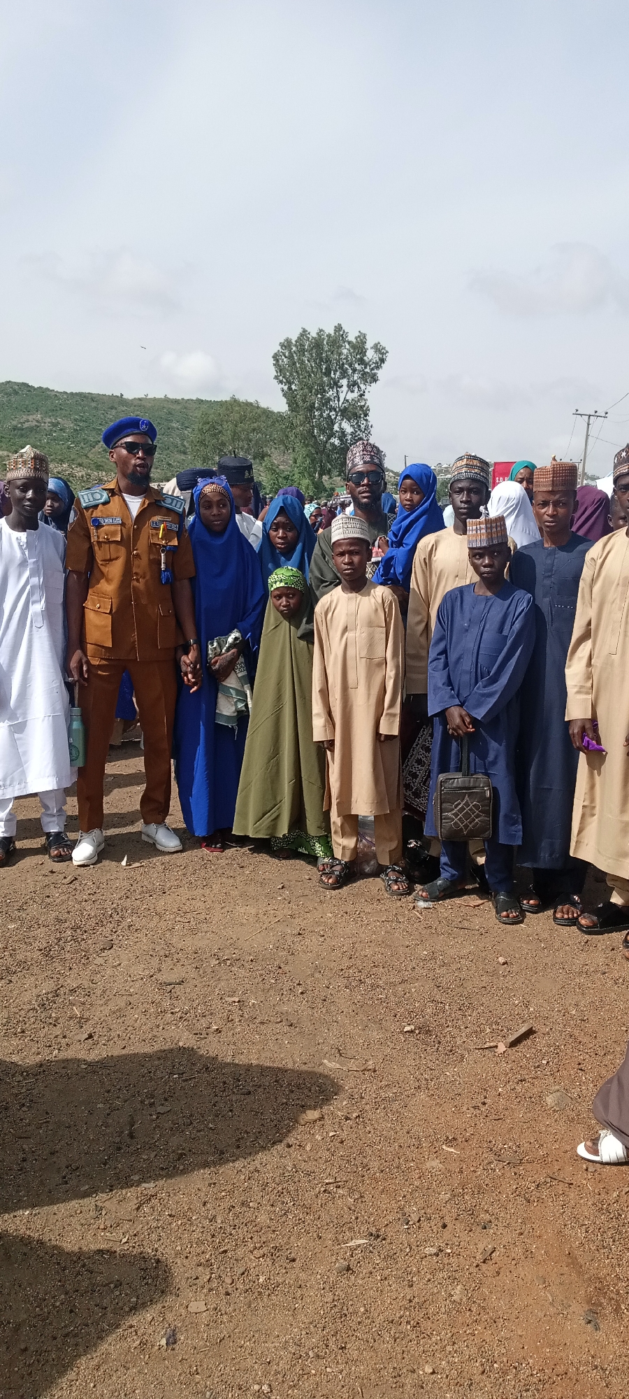 A group of people stand together in an outdoor area in Naraguta Hausawa, Nigeria. The ground is dirt and gravel. In the background, there is a hill with sparse vegetation and a partly cloudy sky. The group includes men, women, and children, most of whom are wearing traditional Islamic clothing. One man stands out due to his formal brown uniform with a blue beret and sunglasses. He holds hands with a woman in a blue headscarf. Several people are wearing traditional Islamic caps. A boy in a blue robe carries a small black patterned bag.