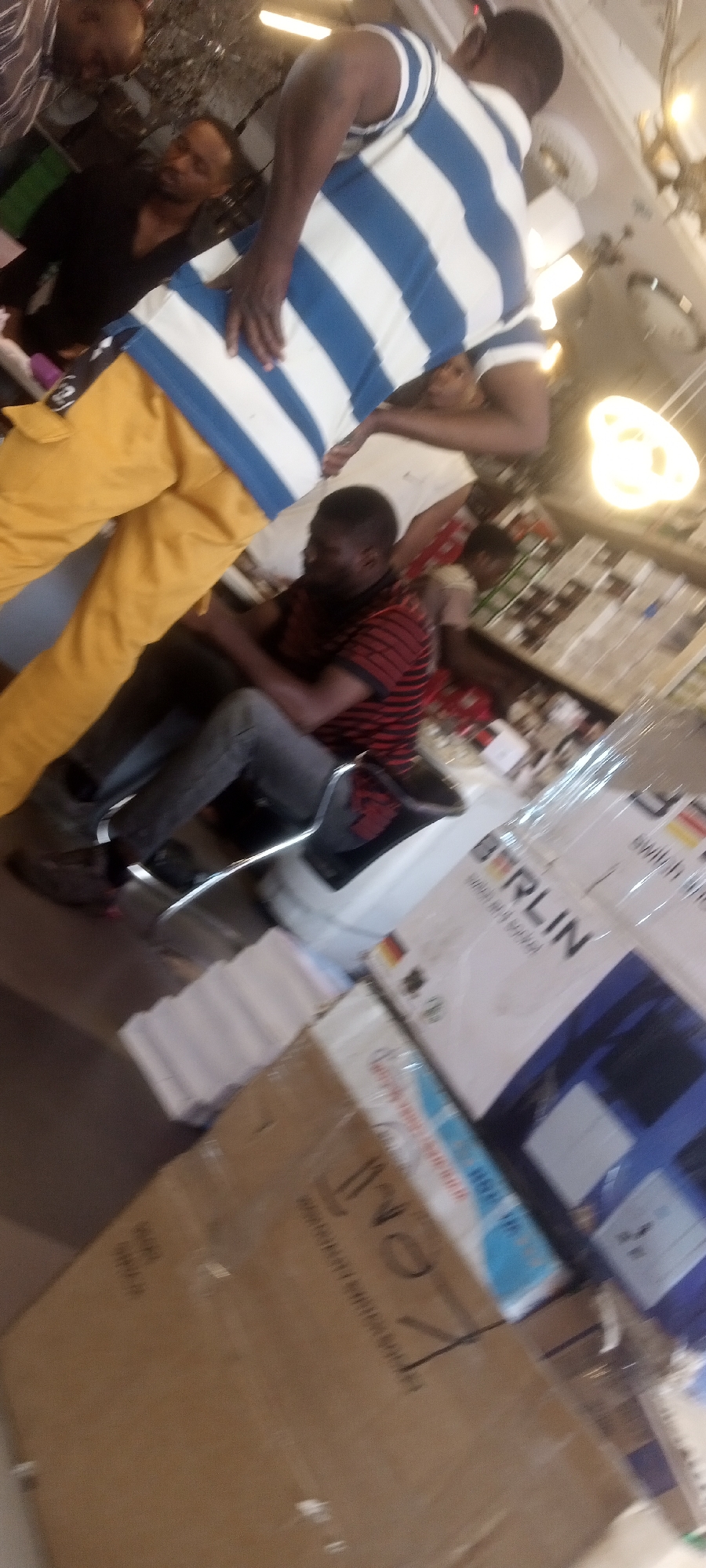 The image appears to be taken inside a shop in the city of Unknown, Unknown. A man wearing yellow pants and a white and blue striped shirt is standing with his hand on his hip. Another man is seated on a chair, wearing a red and black striped shirt and gray jeans. Several boxes are visible in the foreground, with the word "BERLIN" printed on one. Other people are standing in the background. The overall environment seems to be a retail setting with goods on display.