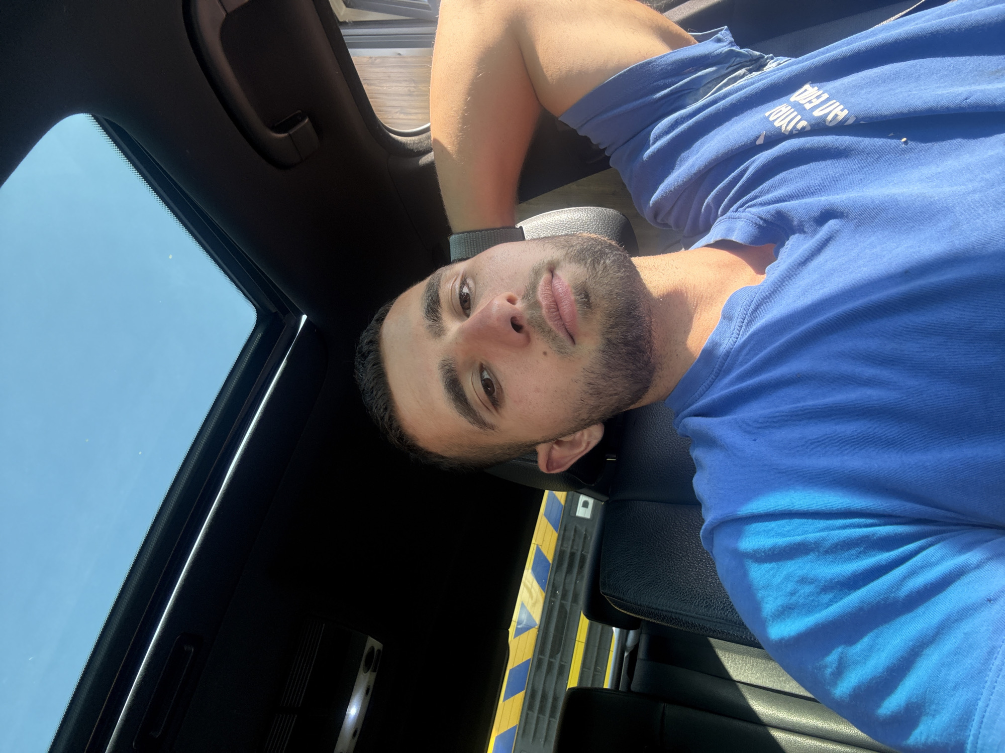 A young man with short dark hair and a beard is sitting in the driver's seat of a car, looking at the camera. He is wearing a blue t-shirt and has his right arm resting behind his head. The car's sunroof is open, revealing a clear blue sky. The interior of the car is visible, including the black leather seats and a section of the dashboard. In the background, there is a yellow and blue striped barrier, possibly from a construction site or road closure. The lighting suggests it is daytime, with sunlight casting shadows on the man and the car's interior. The image does not provide any specific details to locate it within the city of Gabrovo, Bulgaria.