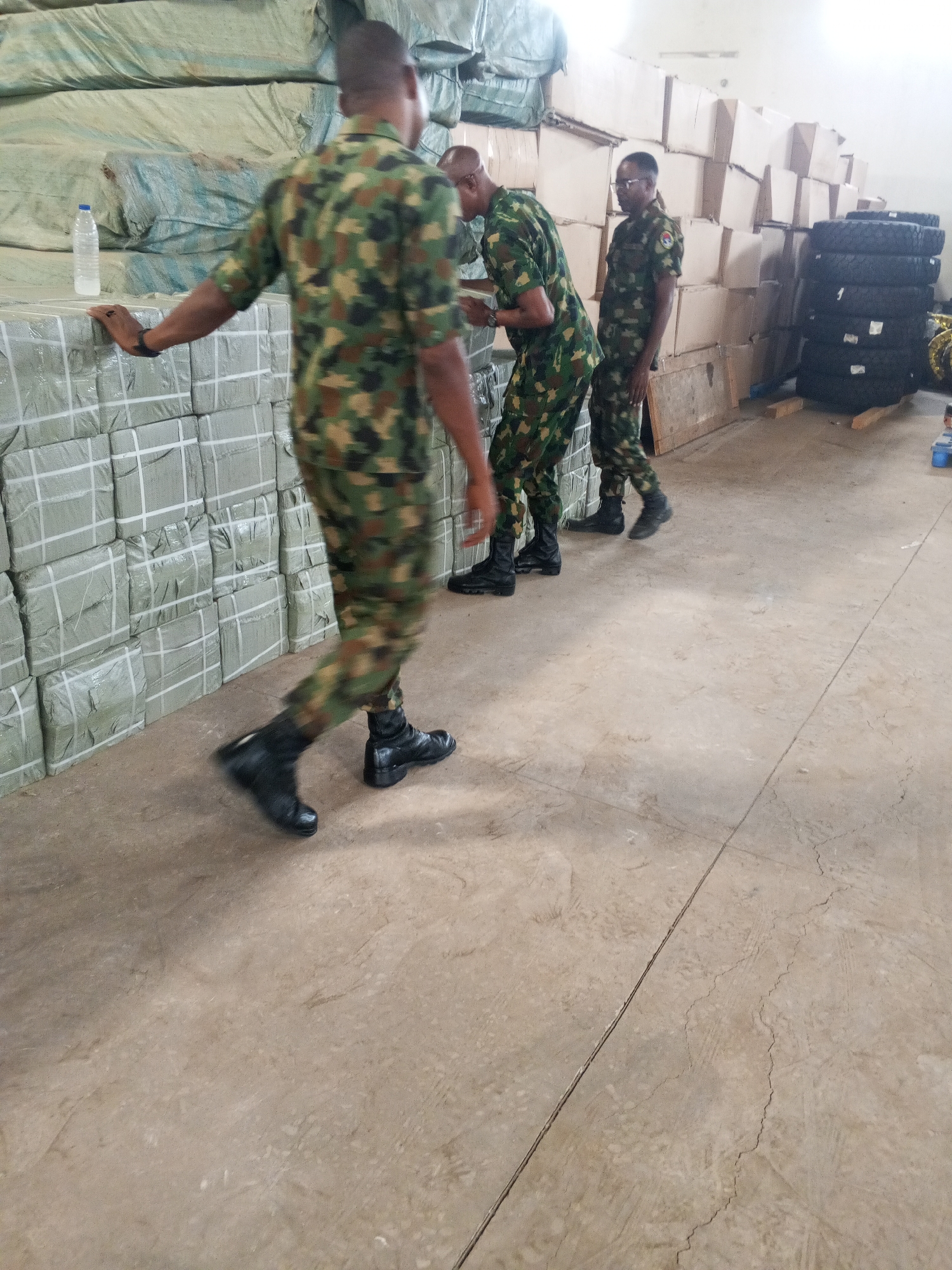 Three individuals in military camouflage uniforms stand in a warehouse. The foreground shows one individual walking towards the camera, with their hand on a stack of packaged goods wrapped in light green material with a white grid pattern. Behind them, two other individuals in similar uniforms are standing near a row of cardboard boxes. To the right, a stack of tires is visible. The floor is a light-colored concrete surface with cracks. A water bottle sits on top of the wrapped packaged goods.