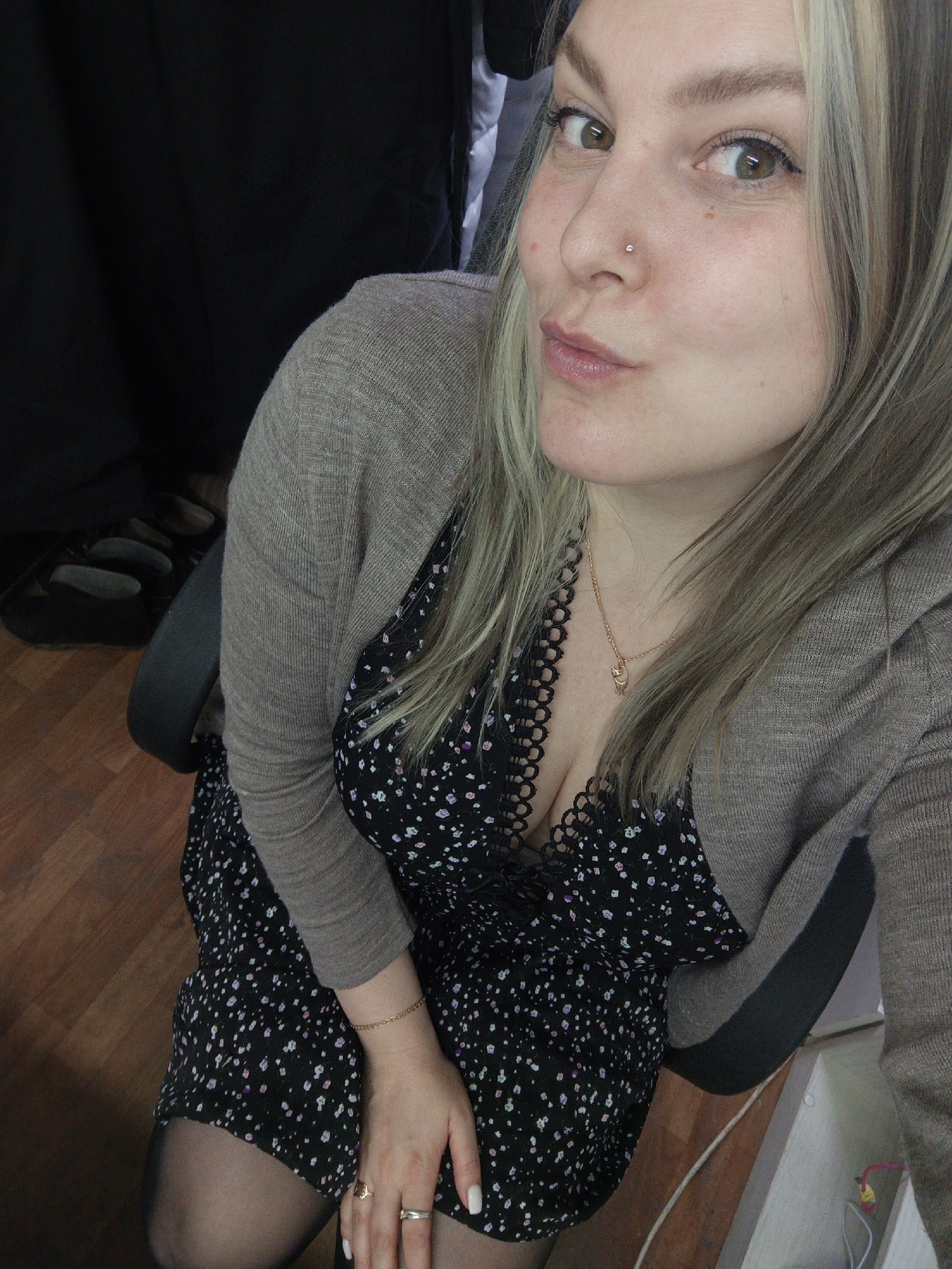 A young woman with light blonde hair and brown eyes poses for a selfie. She is wearing a black floral dress, a light brown cardigan, and black tights. A gold necklace and rings are visible. She is sitting in a black office chair in front of a wooden floor. A black curtain is visible in the background. The location is not discernible from the image.