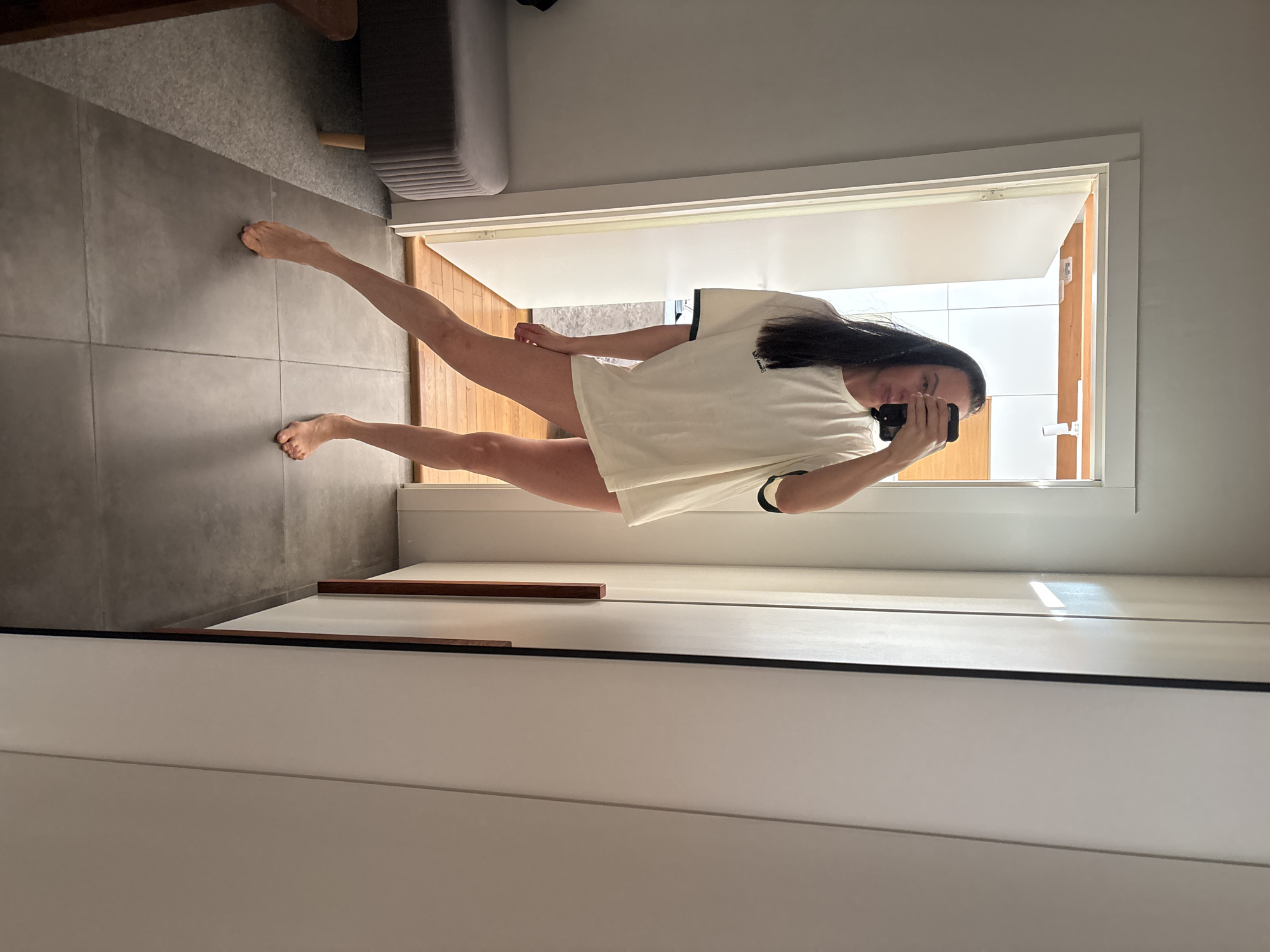 A female individual is observed in a mirror selfie within an interior space in Chaikovsky, Russia. She stands barefoot on dark gray square tiles, with her left leg extended forward on the ball of her foot and her right foot flat. She wears a loose-fitting, light-colored t-shirt with dark trim on the sleeves, with no lower garments visible. Her long dark hair hangs over her shoulders, and she holds a black smartphone to her face, obscuring part of her reflection. To the left, a portion of a dark gray upholstered bench with wooden legs is visible on a lighter gray carpeted floor. Behind the individual, an open white door reveals an adjacent room with wooden flooring and light-colored walls. On the right side of the frame, a floor-to-ceiling white wardrobe or cabinet with dark vertical accents is present.
