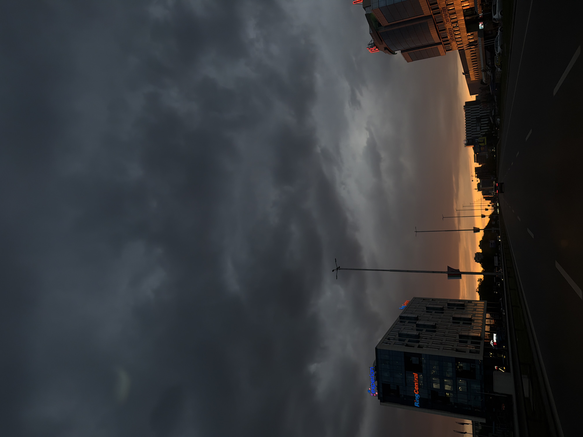 The image shows a landscape in Sofia, Bulgaria. In the foreground, there is a view of a highway, with a building located on the right side. The building has a sign saying "Ring Central" on the top. The sky is filled with dark clouds. There are street lights extending along the road. Other buildings can be seen along the horizon. The overall color palette of the image shifts from dark grey on the left to orange and black on the right, indicating a sunset or sunrise occurring behind the buildings on the right. The image is taken at an angle rotated approximately 90 degrees counterclockwise.