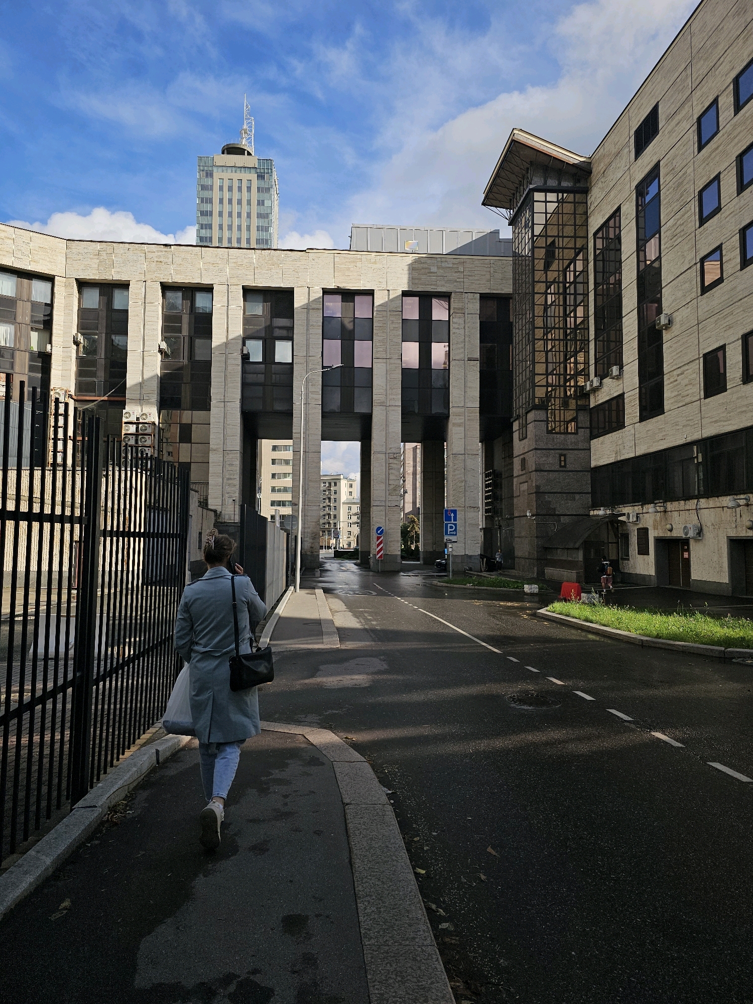 A woman walks down a sidewalk next to a road in front of a modern building with a stone facade. The building has a large arched entrance. A taller, glass skyscraper with an antenna is visible in the distance. The sky is blue with white clouds. The woman is wearing a light blue coat, jeans, and sneakers, and is carrying a black bag and a white plastic bag. The scene appears to be in Moscow, Russia, as indicated by the architecture and street signs.