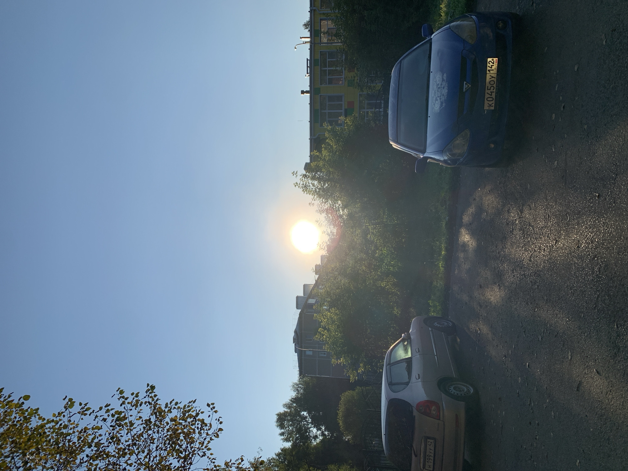 The image depicts an outdoor scene under a clear blue sky with the sun prominently positioned in the upper center, casting a bright glow and lens flare. In the foreground, two cars are parked on an asphalt surface. On the left is a light-colored hatchback with its rear visible, showing a license plate that reads "H 199YY 42". On the right is a dark blue sedan facing forward, its license plate displaying "K0450Y 142" with "RUS" below it. Behind the cars, lush green trees and bushes create a dense foliage. Further in the background, there are residential buildings. The building directly behind the blue car appears to be a colorful school or kindergarten, with yellow walls and multi-colored squares (green, red, blue) on its facade, and large windows. To the left of this building, another, plainer gray residential building with visible windows is partially obscured by trees. The overall impression is a residential area in Prokopyevsk, Russia, during daylight hours, likely late afternoon given the low angle of the sun. The scene is static, with no people or active events visible.