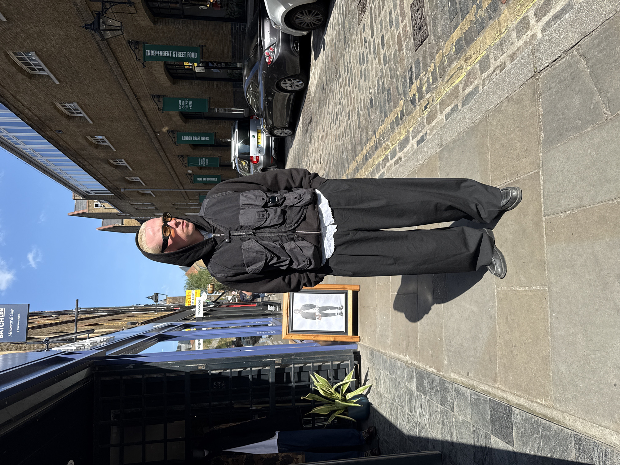 The image captures a sunlit street scene in London, United Kingdom, featuring a person standing on a stone sidewalk. The individual has light-colored hair and wears sunglasses, a black hooded jacket with a multi-pocketed utility vest over a white top, black wide-leg trousers, and black shoes. Their shadow stretches prominently on the pavement to their left, indicating a bright, clear day.
Behind the person, a cobblestone street with a yellow line is visible, where several cars are parked, one displaying a "UK" license plate. Old brick buildings line the street on the right, adorned with various commercial signs. Text from these signs includes "INDEPENDENT STREET FOOD," "LONDON CRAFT BEERS," "WINE AND COCKTAILS," and "seven dials market KERB," suggesting a bustling market or food district. Another sign reads "BATCH LON Menswear & Cafe SHORTS GARDENS," pinpointing a specific location.
In the foreground, a storefront with dark panels and a glass canopy reflects the clear blue sky. A potted snake plant sits on the ground beside a framed picture displayed on the sidewalk, which depicts a person in similar attire. Other visible texts include "TO LET ARL & COUTTS," "eipo," and "topologie." A metal plate marked "METER" is embedded in the cobblestones. The scene portrays a typical urban environment with retail and food establishments under bright daytime conditions.