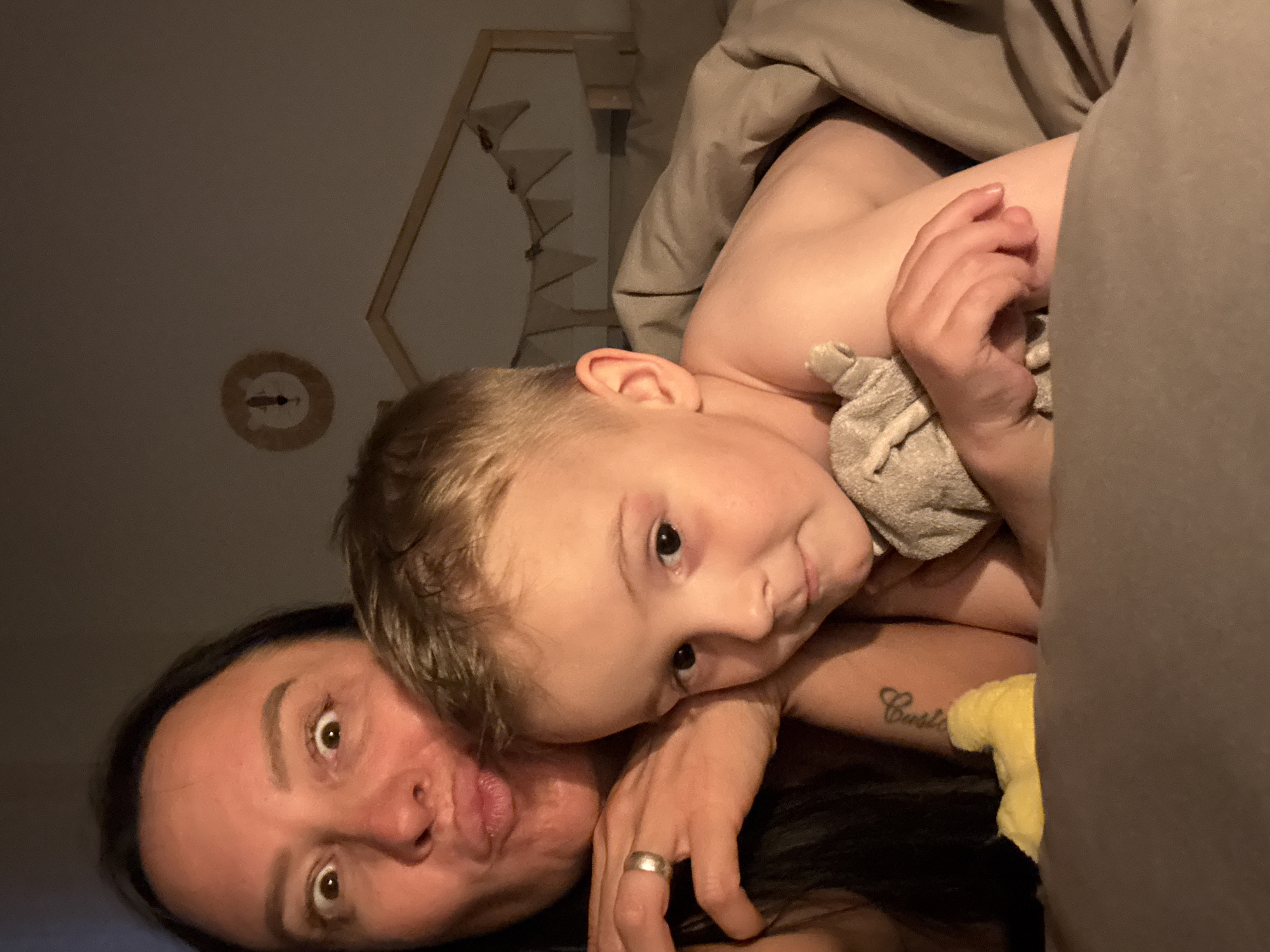 The image depicts a woman and a young child in a bed covered with a gray blanket. The woman is lying on her back and is looking up at the camera, making a puckered expression. The child is lying on the woman, also looking at the camera, holding a gray cloth. The woman is wearing a wedding ring on her finger. Part of a tattoo is visible on her arm. The background shows a light-colored wall with decorative shelves and a light fixture. The image appears to have been taken indoors. No evidence indicates the location of Chaikovsky, Russia.
