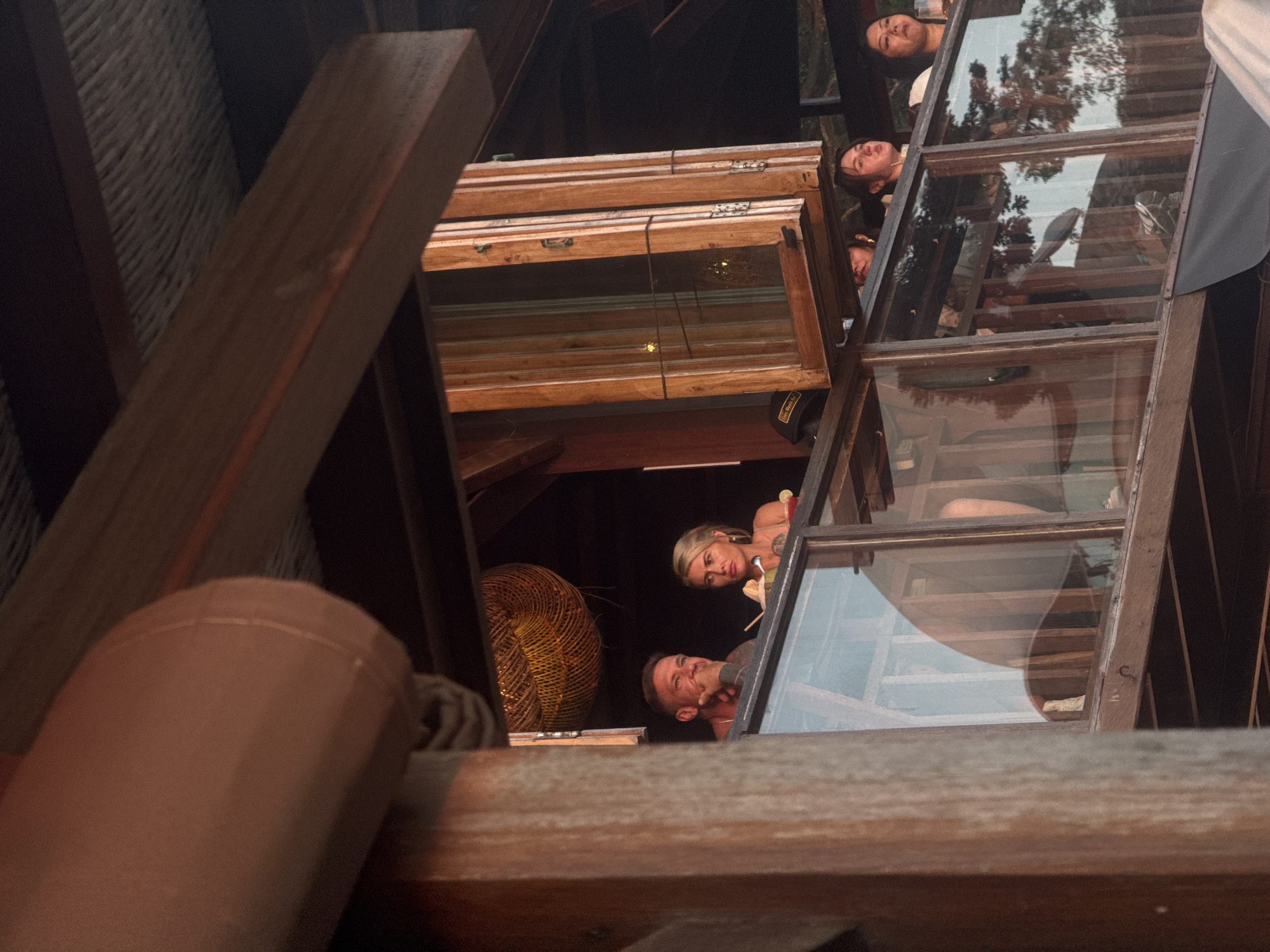In Kuta Selatan, Indonesia, an outdoor restaurant setting is depicted. The image focuses on a couple standing on an upper level balcony, separated by a glass partition and a wooden railing. The man, on the left, has short, dark hair and is wearing a dark shirt with tattoos visible on his arms and chest. He is leaning his chin on his hand, appearing to be in thought. To his right, a woman with blonde hair pulled back and wearing a light-colored top holds a drink in her hand. She is looking towards her right. A woven light fixture is visible in the background. Below them, several other people are seen on a lower level, also behind glass. The architecture features prominent wooden beams and structures, with a thatched-looking ceiling visible in the upper part of the image. The overall impression is a casual, outdoor dining environment with a view of lush greenery reflected in the glass.
