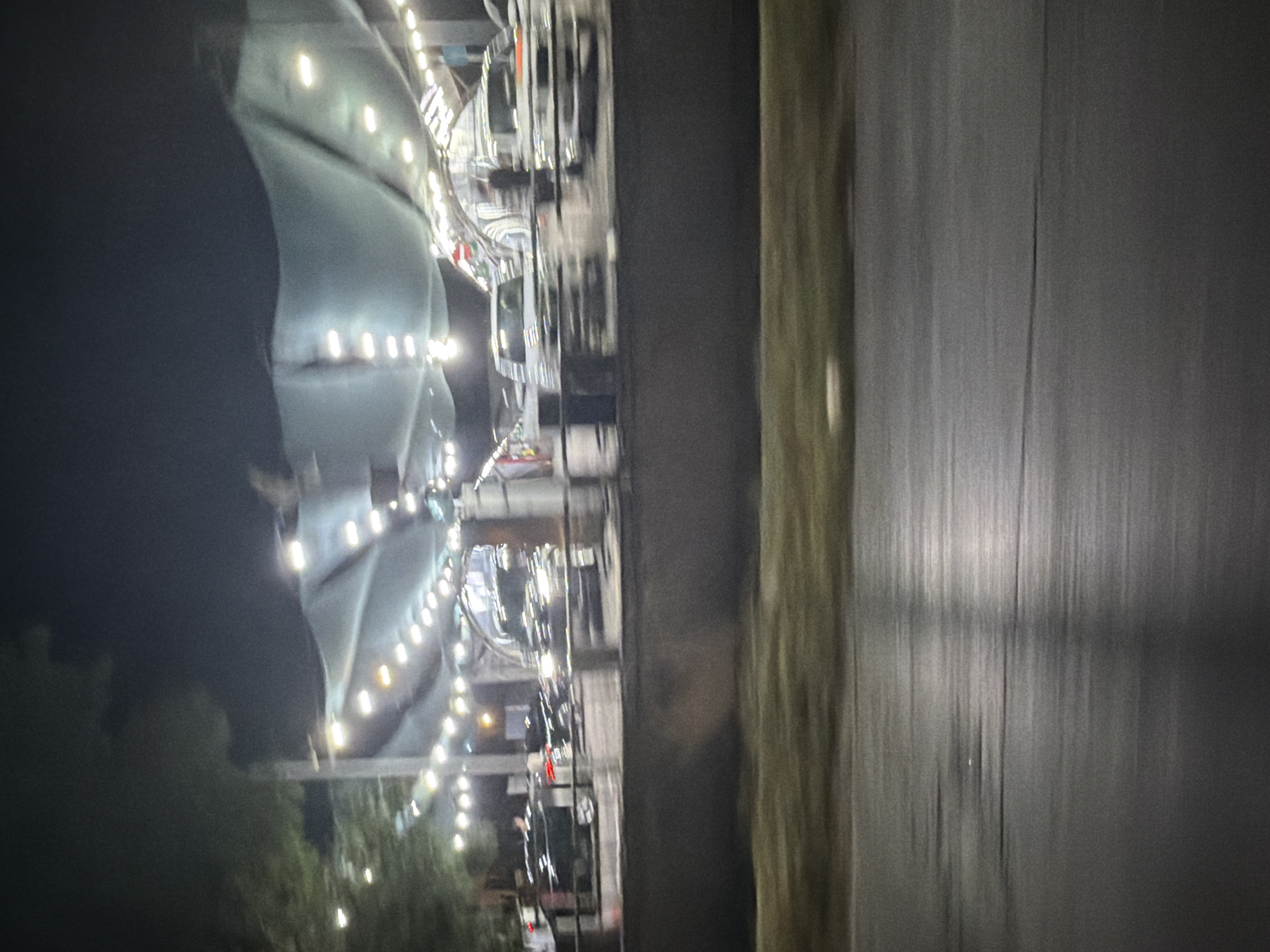 The image shows a blurry nighttime view of a car wash. Several vehicles are visible under a large, illuminated canopy. The cars appear to be clean and in various colors, predominantly black and white. The lighting is bright, with strings of lights illuminating the area. The car wash is situated in what appears to be an urban setting, with a concrete barrier and some vegetation visible in the foreground. The overall impression is one of a well-lit, active car wash at night. The crops do not provide any specific details that would indicate the location in Tashkent, Uzbekistan.