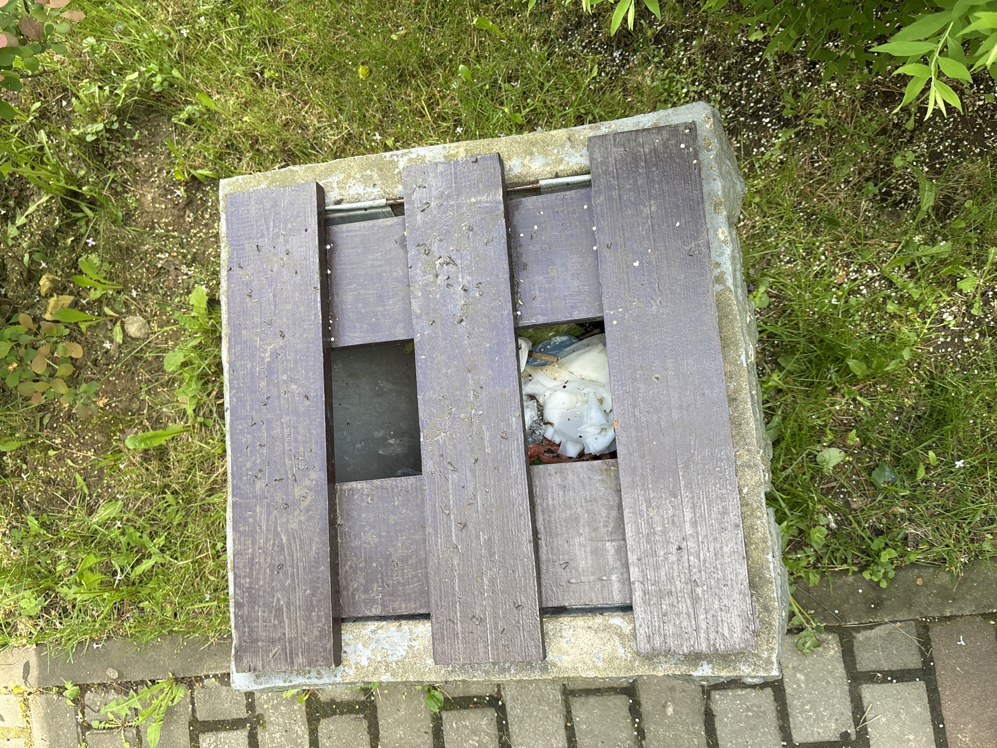 A top-down shot shows a gray, partially-filled trash receptacle in a park-like setting. The receptacle is square, constructed with wood slats spaced horizontally over a concrete base. Litter, including white and reddish materials, is visible inside. The container is situated on a brick surface adjacent to grassy areas with small plants and leaves scattered around. The environment suggests an outdoor public space with landscaping. There are no people visible in the frame. The photo was likely taken in Saint Petersburg, Russia.