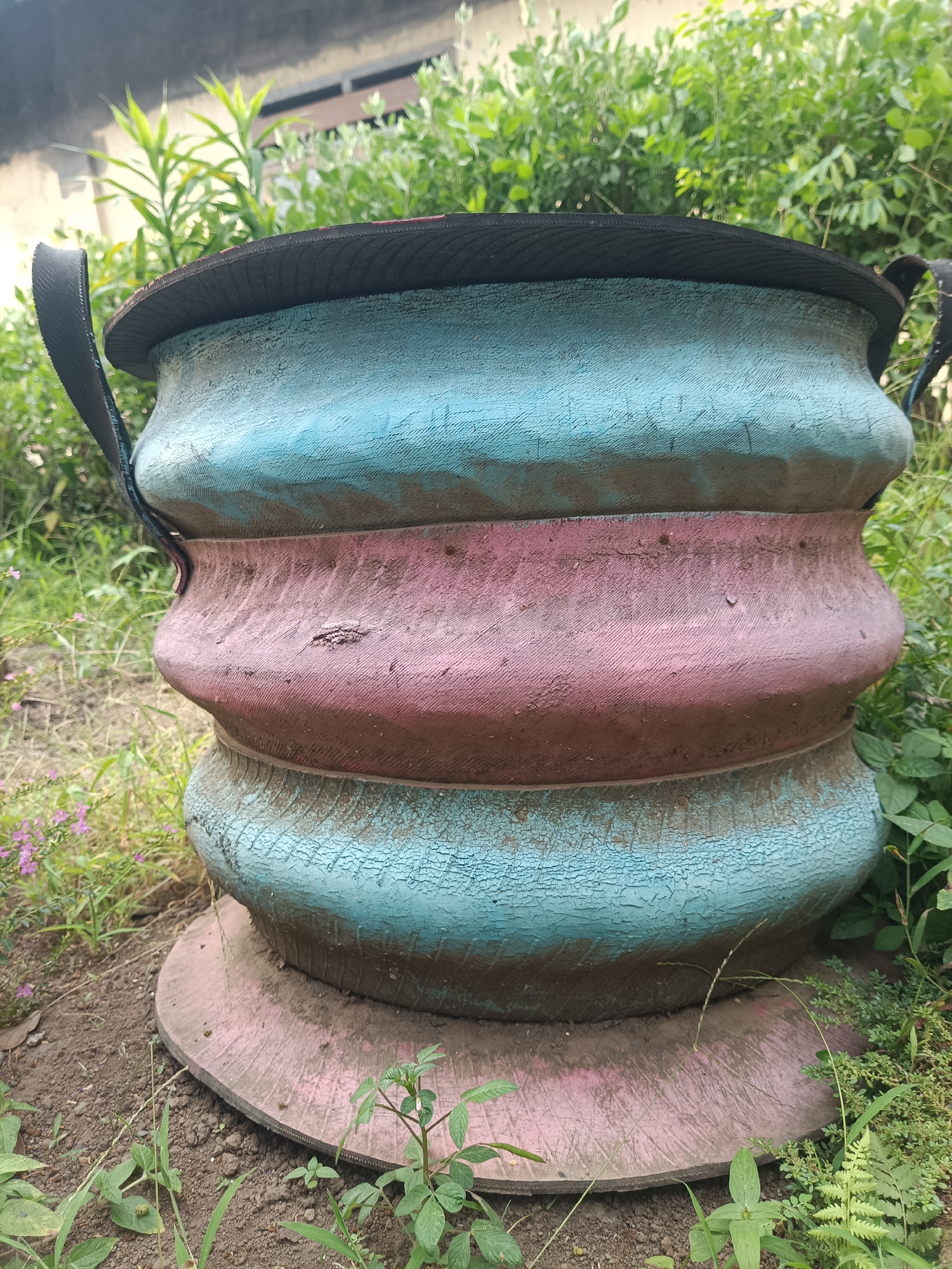 The image shows a large, multi-tiered planter made from painted tires. The planter is predominantly teal and pink, with signs of wear and cracking in the paint. It has two black handles visible on opposite sides. The planter sits on a circular, flat base, also painted pink, which is resting on the ground. The surrounding environment is overgrown with various green plants and some dry soil, suggesting an outdoor setting. In the background, there is some foliage and a hint of a building or wall. This scene is located in Jember, Indonesia.