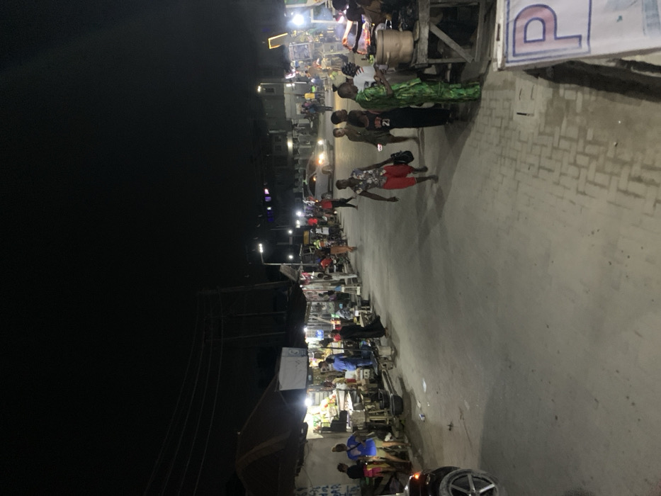 The image depicts a nighttime street scene in Iranla, Nigeria, characterized by artificial illumination. The environment consists of a paved road with an adjacent sidewalk on the right, bordered by multiple makeshift market stalls and permanent structures on both sides. Overhead utility lines are visible against the dark sky.
Approximately 20-30 individuals are present. On the left, several people congregate near stalls displaying various goods under bright white lights. A dark SUV is partially visible in the foreground on the left. In the mid-ground, a person wearing a floral shirt and red shorts walks towards the viewer, holding a dark bag. Other individuals are walking along the road and congregating near stalls on the right side. A light-colored sedan is visible in the distance, heading away from the viewer.
On the right side, multiple stalls are active, with individuals standing, interacting, and preparing items. One stall in the foreground features a large metal pot on a wooden stand. A partially visible blue and white sign with the letter "P" is in the lower right corner. The ground shows some scattered debris.