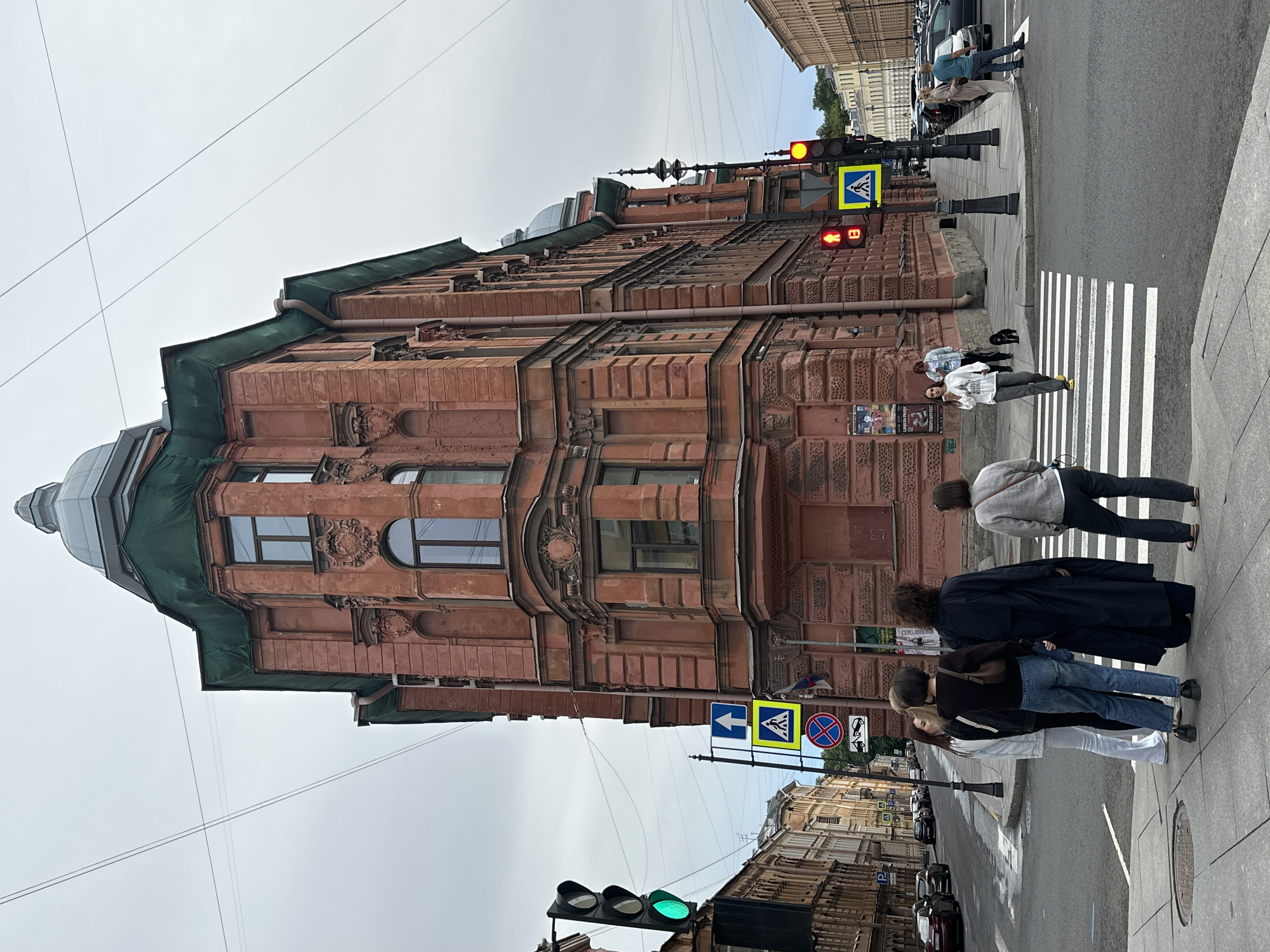 This image captures an overcast day in Saint Petersburg, Russia, likely near the intersection of Nevsky Prospekt and the Moika River embankment. The focal point is a tall, reddish-brown brick building with ornate architectural details and a green temporary covering on its upper sections, suggesting ongoing renovation or repairs. The building's distinctive corner design and multi-story bay windows are prominent.
In the foreground, a group of five individuals, appearing to be tourists or locals, stand on a pedestrian crosswalk, waiting to cross the street. They are casually dressed. One person in the group is taking a photograph of the building. A woman walks across the crosswalk towards the building, accompanied by a small black dog. Another woman, wearing a white long-sleeved shirt and dark pants, walks across the street.
Traffic lights are visible, with one displaying a green signal for vehicles and another showing a red pedestrian signal. Several street signs are also present, including a pedestrian crossing sign and a traffic prohibition sign. The street is paved with asphalt, and the sidewalks are made of granite slabs. Cars are parked along the street and driving in the background. The overall atmosphere is urban, with a mix of historical architecture and modern city life.