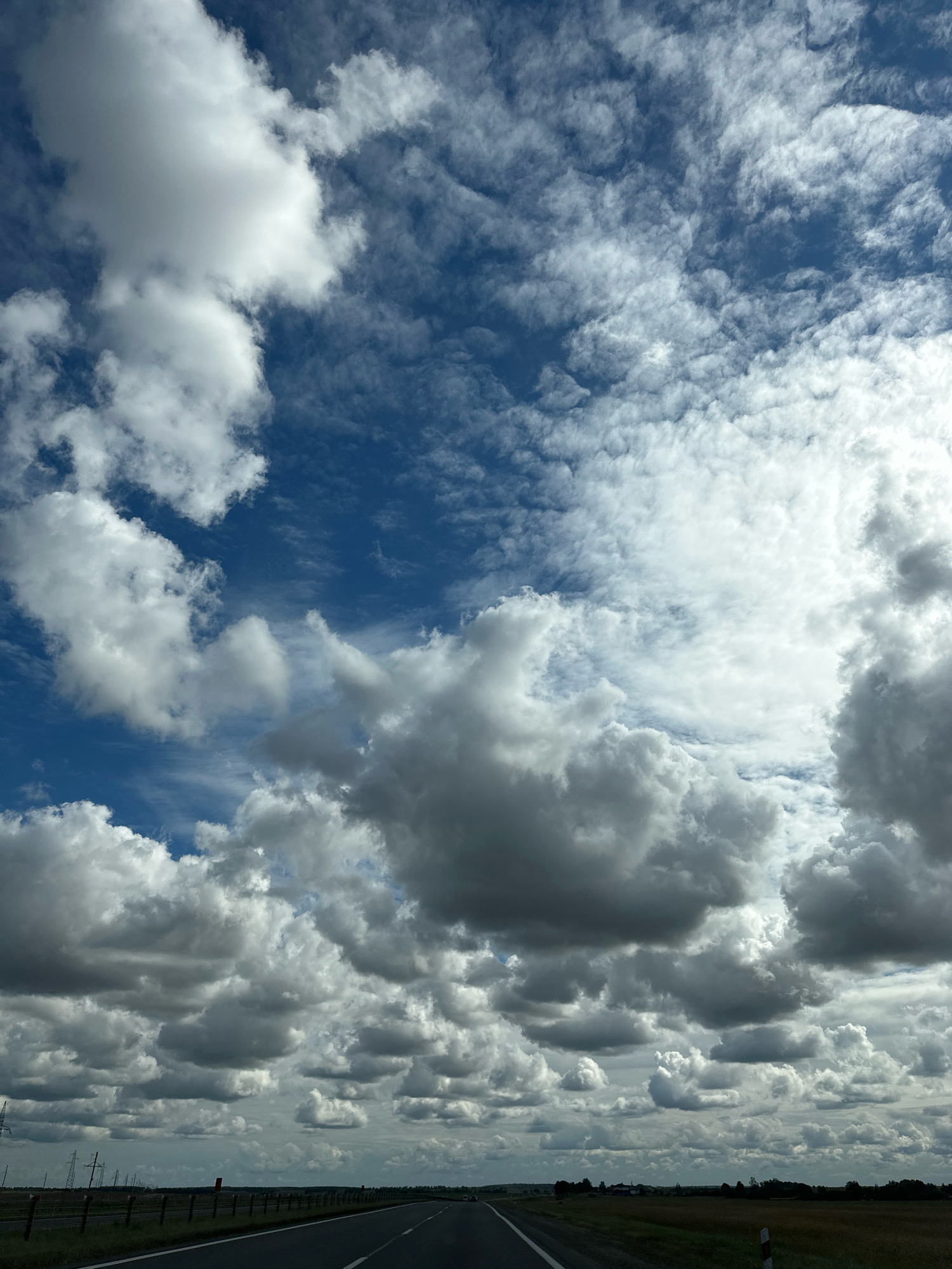 The image depicts a landscape viewed from a road, possibly a highway, as indicated by the paved surface with white lane markings and a shoulder. The sky is filled with a dramatic display of clouds, ranging from fluffy white cumulus to darker, more ominous formations. The ground level shows a flat, rural terrain with fields of vegetation, some appearing dry and harvested, bordering the road. A fence runs along the side of the road, and in the distance, there are signs of human infrastructure, including power lines and what appear to be utility poles. The overall impression is one of natural beauty and open space, under a dynamic sky. The location is described as Блонскі сельскі Савет, Belarus, which is an administrative division within the country.