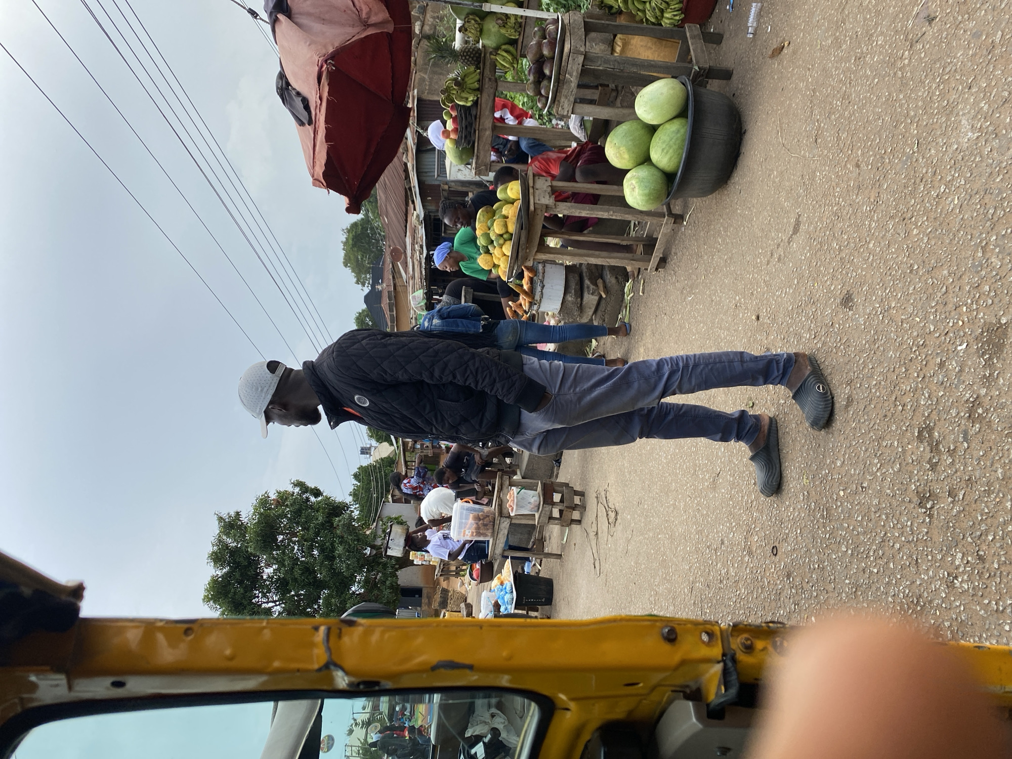 In Bukuru, Nigeria, on a cloudy day, a man wearing a gray cap, a black quilted jacket, and gray pants stands on a dusty street. He is looking to his right. In the foreground to the left, the yellow side of a motorized tricycle is visible. Behind the man, a vibrant market scene unfolds. Stalls are laden with fresh produce, including watermelons, bananas, pineapples, avocados, and piles of citrus fruits. Several people are engaged in market activities, with some tending to the stalls and others browsing. The background shows several buildings with corrugated metal roofs, indicating a dense urban or semi-urban setting. Power lines are visible crisscrossing the sky. The overall impression is that of a busy, local marketplace.