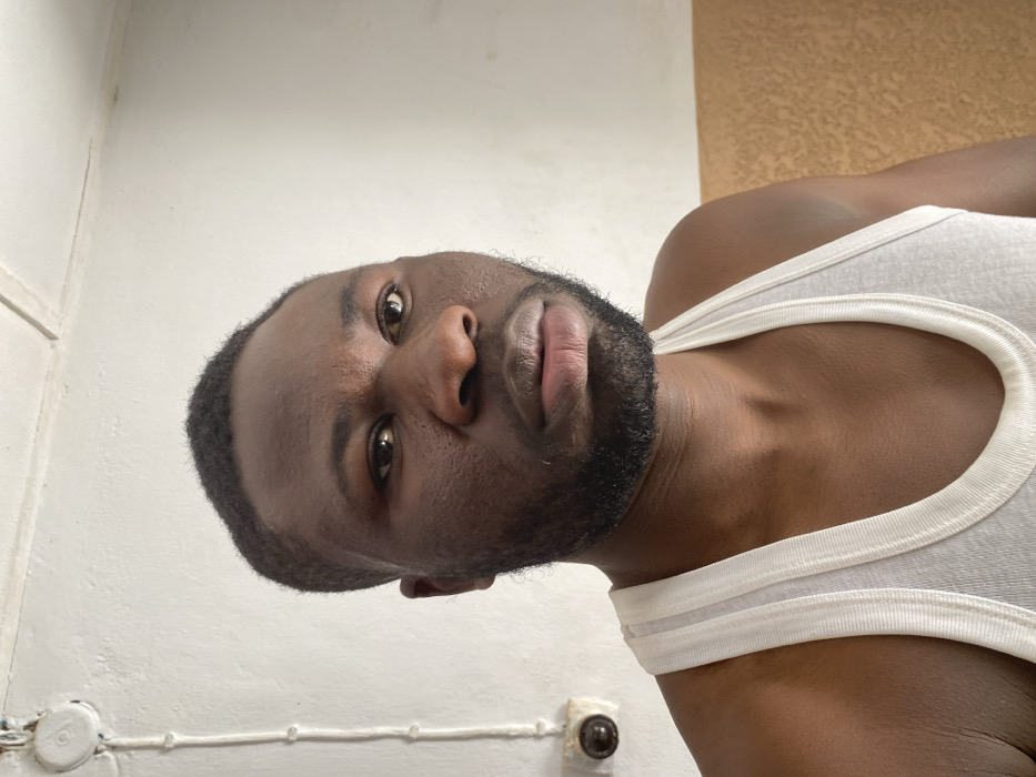 The depicted location is identified as Udu, Nigeria. The image shows a close-up, head-and-shoulders shot of an adult male with dark skin, dark short hair, and a short beard and mustache. He is wearing a white ribbed tank top and looks directly at the viewer with a neutral expression, his head slightly tilted. The background consists of a plain white wall. On the left side of the wall, a white electrical conduit runs vertically, secured by clips, with a round, dark brown electrical component, possibly a switch, attached to it at shoulder height. The lower right portion of the wall indicates a transition to a tan or light brown colored section.