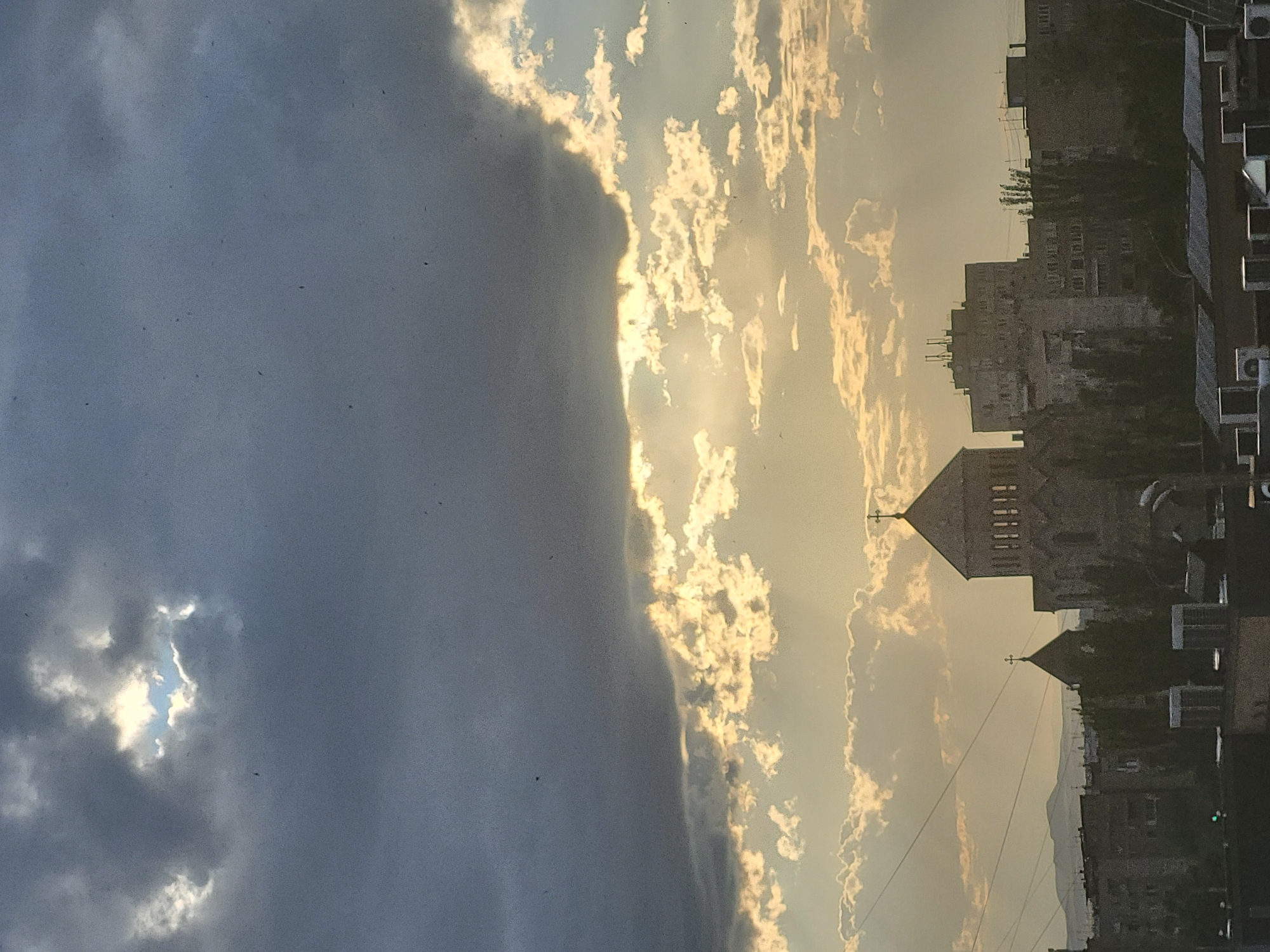 The image shows an urban scene in Yerevan, Armenia, featuring a sky divided between dark, ominous clouds on the left and lighter, sun-illuminated clouds on the right. A church with a prominent cross atop its tower stands amidst buildings, suggesting a cityscape. Power lines are visible in the sky, adding to the urban context.