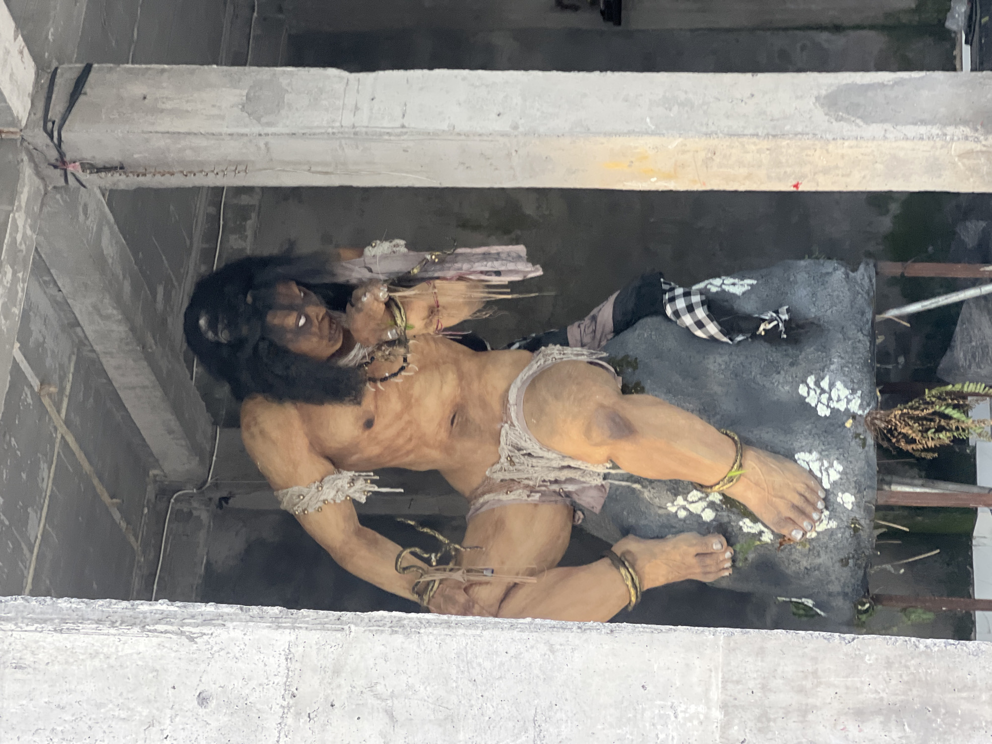 The image shows a large, detailed statue of a dark-haired male figure lying on a gray, textured surface. The statue appears to be made of a material resembling clay or resin and is predominantly tan, with darker accents around the hair and facial features. The figure is partially draped in rough fabric, including a white and black checkered cloth.
The statue is positioned within a gray, concrete structure, possibly the interior of a building or enclosure under construction. The view is from above, looking down into the space where the statue is placed. The concrete structure consists of horizontal beams or ledges. Based on context, this is likely a religious statue in Celuk, Indonesia.