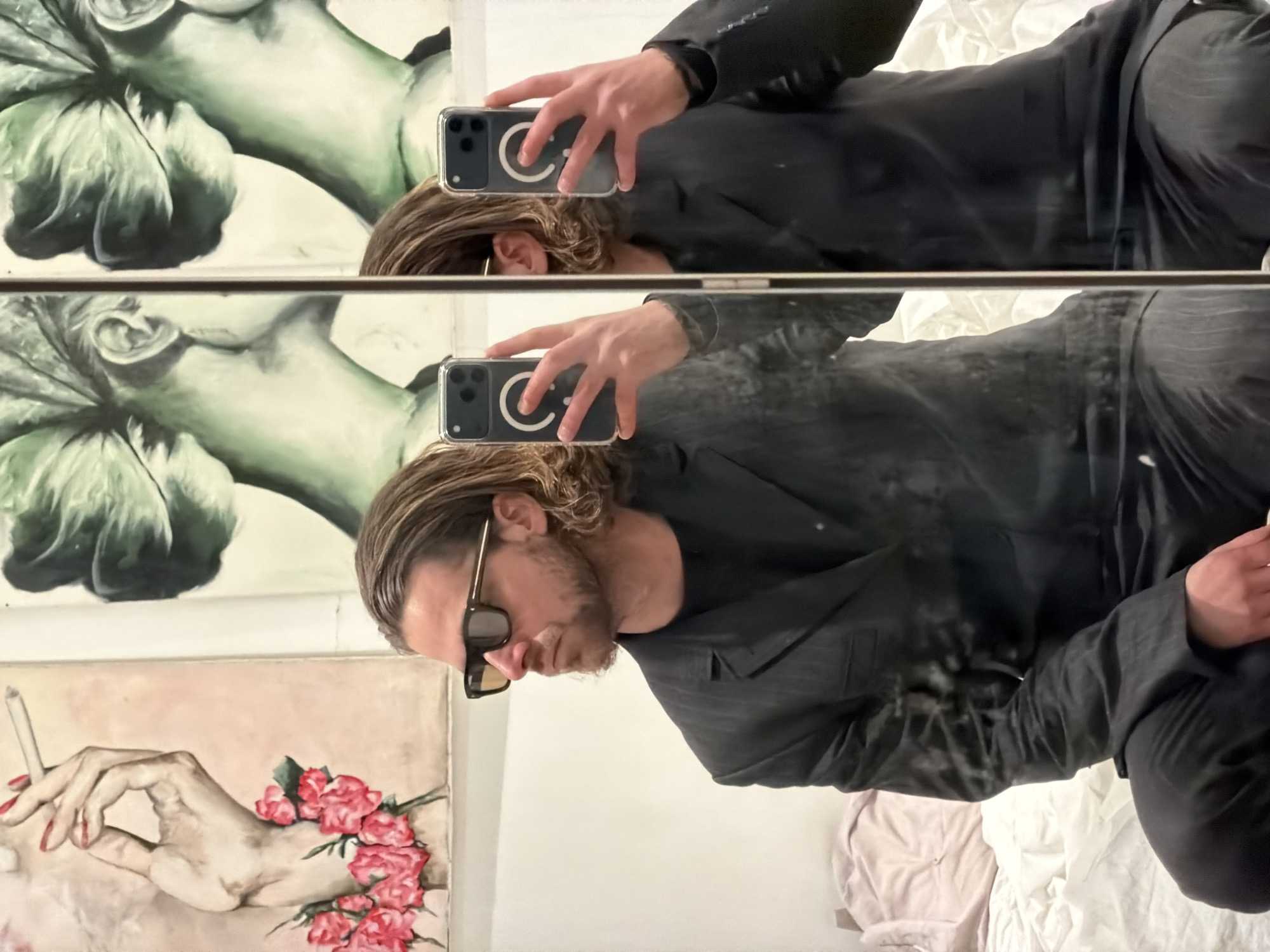 An adult male is captured in a mirror selfie within an indoor room in Paris, France. He is seated on a rumpled bed with light-colored and pink bedding. The subject possesses medium-length, wavy light brown hair, a beard, and wears dark rectangular sunglasses with gold-colored temple accents. He is dressed in a dark, possibly pinstriped, suit jacket over a dark shirt and matching dark trousers. His left hand is raised, holding a smartphone in a clear case with a visible circular magnetic charging ring, pointed towards the mirror to capture the image. His gaze is directed to his left. Behind him, on a light-colored wall, two paintings are visible: one depicts a pale hand with red nail polish holding a cigarette, adorned with red roses; the other shows a stylized green head or bust. A vertical mirror frame divides the image, showcasing the primary reflection and a secondary, slightly offset reflection.