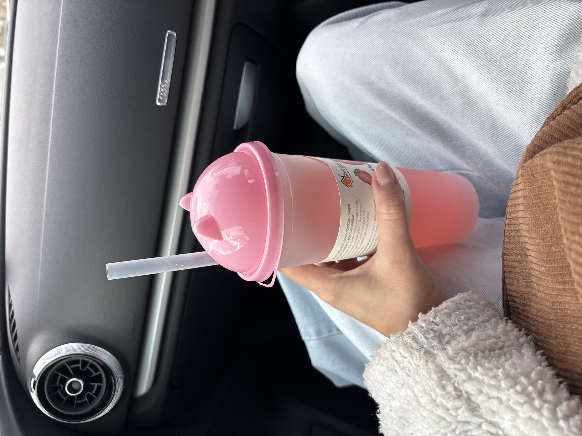 The image captures a person holding a pink, translucent drink cup with a straw while seated inside a car, likely in Krasnoyarsk, Russia. The setting appears to be daytime. The person wears light blue denim jeans and a brown, textured jacket with a fluffy, light-colored sleeve cuff. Their right hand, adorned with neatly manicured nails, holds the drink cup.
The cup contains a pink liquid and has a matching pink lid with small, ear-like protrusions. A clear straw is inserted into the lid. A label on the cup, partially visible, displays text in Cyrillic. OCR analysis reveals details about the drink, including "non-alcoholic juice-containing drink with strawberry flavor," mentions of concentrated strawberry juice, food additives like citric acid and Acesulfame potassium E950, and food dyes. Storage instructions to "store in a dry and cool place" are also visible.
The car's interior features include a dark grey door panel with a silver, elongated door handle/trim piece and a distinctive circular air vent with a knurled silver ring, characteristic of Audi vehicles. The overall scene suggests a casual moment, perhaps commuting or traveling, with the person enjoying a flavored drink.