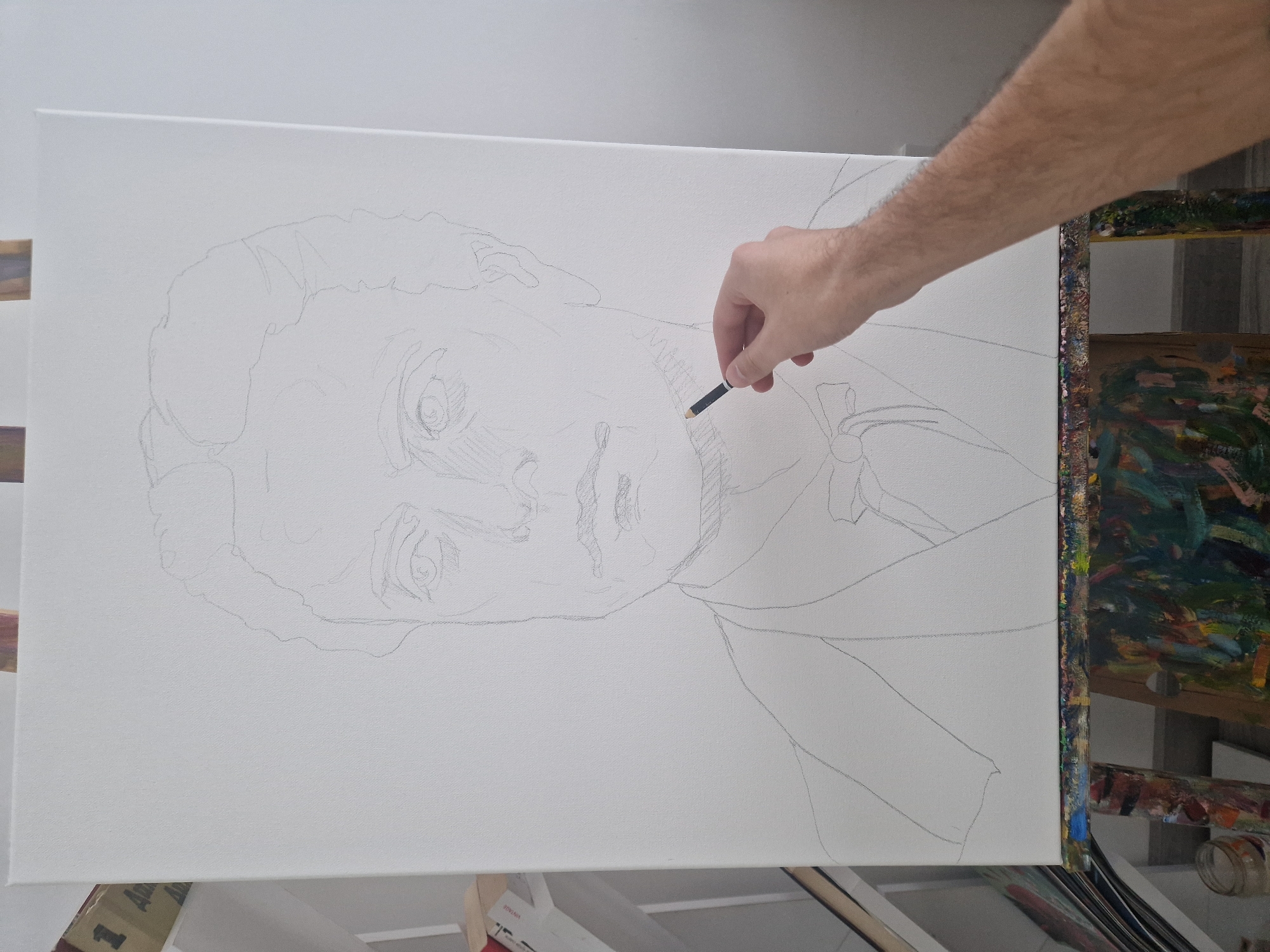 A close-up indoor shot shows a partially completed pencil sketch of a person's face on a rectangular white canvas mounted on an easel. The sketch depicts a man's head and upper torso. An arm and hand holding a short pencil are visible in the upper right of the image; the person is likely sketching the artwork. The easel's edge is visible to the right of the canvas, showing dried paint. The city and specific location are unknown.
