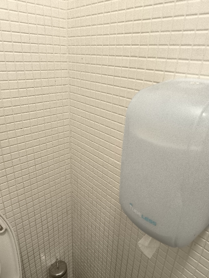 The image presents a static interior view of a clean restroom corner in Palaio Faliro, Greece. The walls are uniformly covered with small, rectangular, light beige or off-white tiles arranged in a grid pattern, creating a textured surface.
A white or light gray wall-mounted hand dryer dominates the right side of the frame. A small, crumpled piece of white paper or tissue hangs from the dispenser slot at its bottom. Near the lower central part of the hand dryer, light blue or teal text reads "Tube Less", accompanied by a subtle, wave-like or abstract logo positioned above the "L" of "Less". The hand dryer's surface has a slight sheen or fine texture.
In the bottom-left corner, a portion of a light-colored toilet lid is visible. Situated on the floor next to the toilet is a metallic, cylindrical toilet brush holder. The lighting throughout the space is even and bright, suggesting indoor artificial illumination. No individuals are present, and no activity is depicted, making it a straightforward architectural or facility shot. The restroom appears well-maintained and utilitarian.