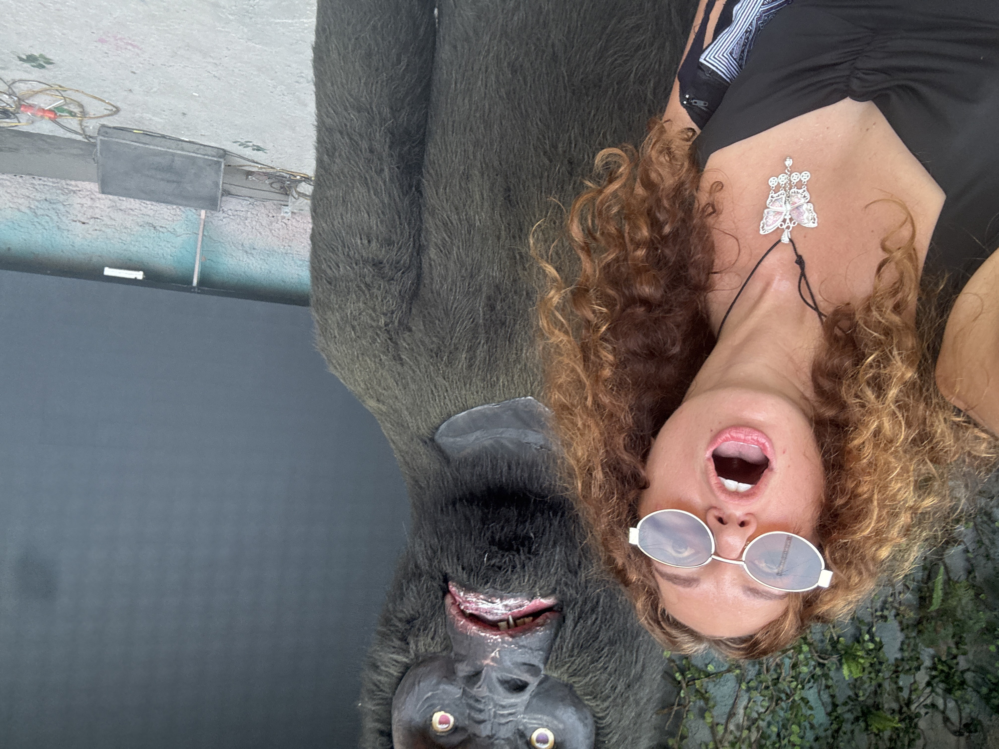 A woman with curly brown hair and sunglasses is taking a selfie in front of a large, furry ape statue. The woman is wearing a black top and a butterfly pendant necklace. The ape statue is dark gray and has realistic features, including fur, eyes, and a mouth with visible teeth. The background appears to be an outdoor setting with some greenery and a dark wall. The ape statue's left arm is extended, and the woman's left arm is visible as she holds the phone to take the picture. The image is taken from a medium shot, with the woman and the ape statue occupying most of the frame.