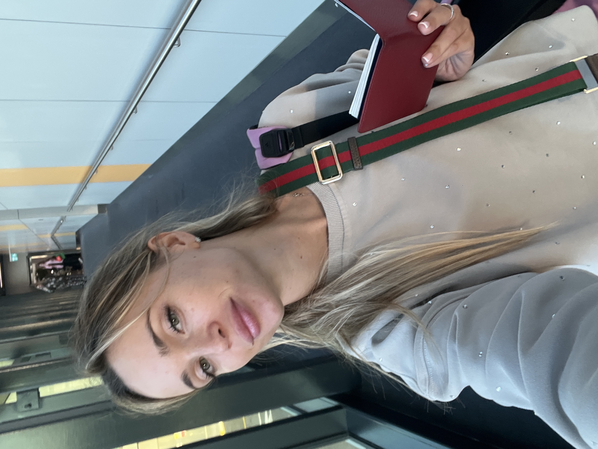 In Arnavutköy, Turkey, a woman with blonde hair and green eyes is taking a selfie in an airport. She is wearing a light-colored sweatshirt adorned with small, sparkling embellishments. A bag with a green and red striped strap and a pink strap is visible over her shoulder. In her right hand, she holds a red book-like object, possibly a passport holder. The background shows an airport corridor with a dark floor and a railing along the right wall, illuminated by natural light from large windows on the left.