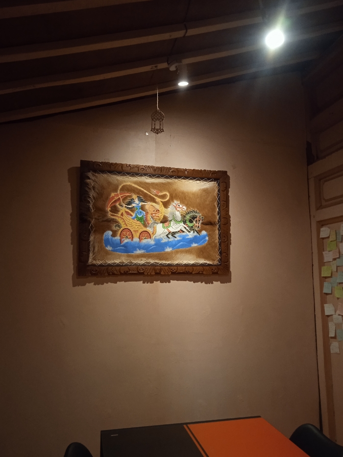 The image shows a close-up of a wall inside a building in Surakarta, Indonesia, located at coordinates -7.587 latitude and 110.823 longitude. The wall is painted a pale beige color and is illuminated by a single light fixture mounted to the ceiling. The fixture is a standard white bulb encased in a circular metal housing. A framed painting hangs on the wall, featuring an image of a carriage pulled by horses. The frame is wooden with a brown stain and a raised border. The image inside the frame appears to be a painted depiction of an elaborate chariot with three figures inside, against a background of a blue sky with white waves. The frame is centered on the wall and hangs slightly off center, approximately one-third of the way down from the top of the wall. The scene is quiet and empty. A table with a black top and orange surface extends out of the frame to the right, with a black chair partially visible at the bottom of the image. The wall to the right is a pale yellow color and is covered with an assortment of small colorful sticky notes. The floor in the space is not visible in the image. The scene is brightly lit and captures the interior of what appears to be a restaurant or dining room.