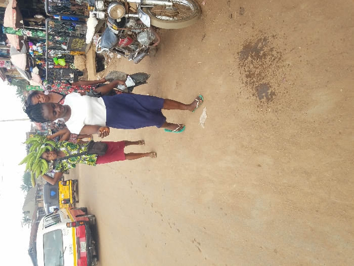 The image depicts a street scene in the city of Nnobi, Nigeria, characterized by an unpaved, light brown dirt road under an overcast sky. Three adult females are walking from left to right across the frame.
In the foreground, a woman wears a white short-sleeved top, a navy blue skirt, and green flip-flops, holding a black plastic bag in her right hand. Her gaze is directed downwards. Behind her, a second woman carries a large, prominent bunch of green plantains on her head. She is attired in a vibrant, patterned short-sleeved top, a red knee-length skirt, and sandals, with a black messenger bag slung over her left shoulder. Partially visible behind the second woman is a third female, wearing a patterned top.
The right side of the street features a row of market stalls or shops with awnings and various goods, fronted by a black metal barrier. Two motorcycles are parked vertically along the right edge of the road. In the left background, a white mini-bus with red and yellow markings is visible, followed by a yellow tricycle (keke napep). Utility poles and overhead wires are present in the distant background. The road surface in the immediate foreground exhibits a dark, damp patch and some scattered debris.