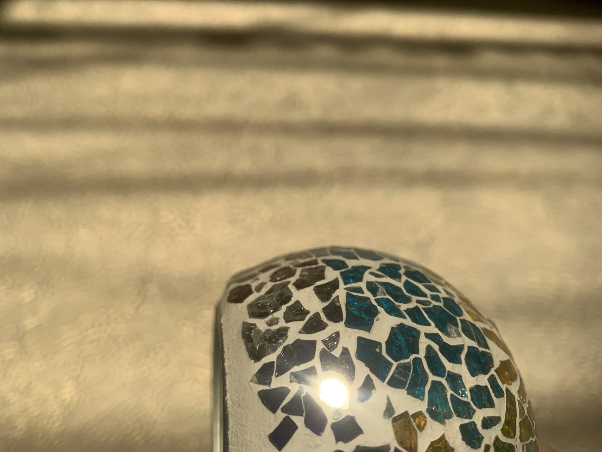 The image features a close-up of a mosaic-covered object, likely a decorative item. The mosaic consists of small, irregularly shaped pieces of glass in various colors, including shades of blue, gray, gold, purple, and green. The pieces are set against a white grout background. A strong light source is reflected off the surface of the mosaic, creating a bright spot. The object has a curved, rounded shape. The background is blurred and appears to be a surface with horizontal lines or patterns. There is no indication of Izhevsk, Russia, or any specific location. No people are visible, and no actions or interactions are depicted.