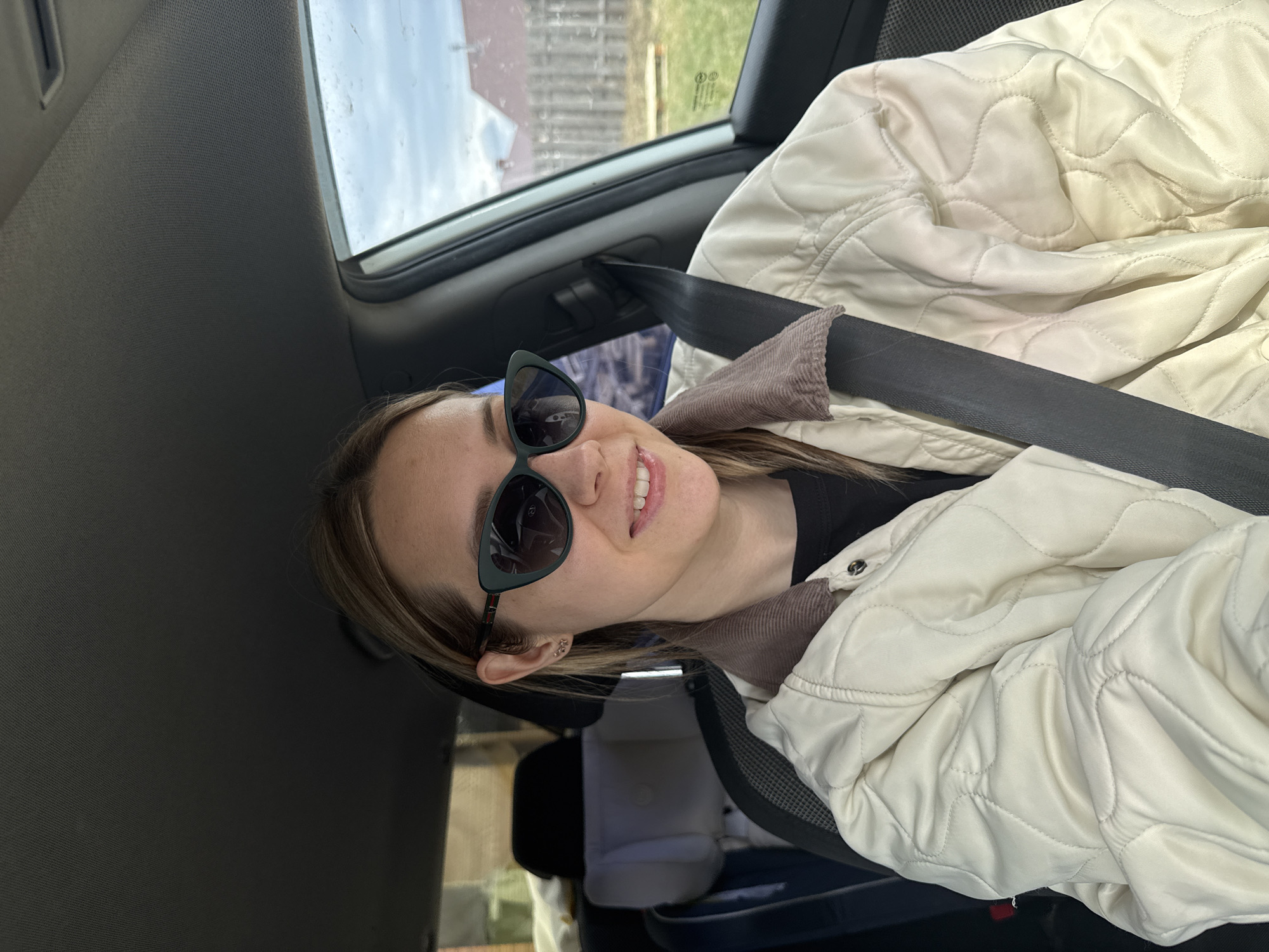 A person is in the driver's seat of a car. The person is wearing sunglasses, a black shirt, and a beige, quilted jacket with a corduroy collar. A seatbelt is fastened across the person's chest. The person is smiling. The background shows a blurred view of a building and vegetation through the car window. Based on the location provided (Chaikovsky, Russia), the architecture and the surrounding environment would be typical of a Russian urban landscape.